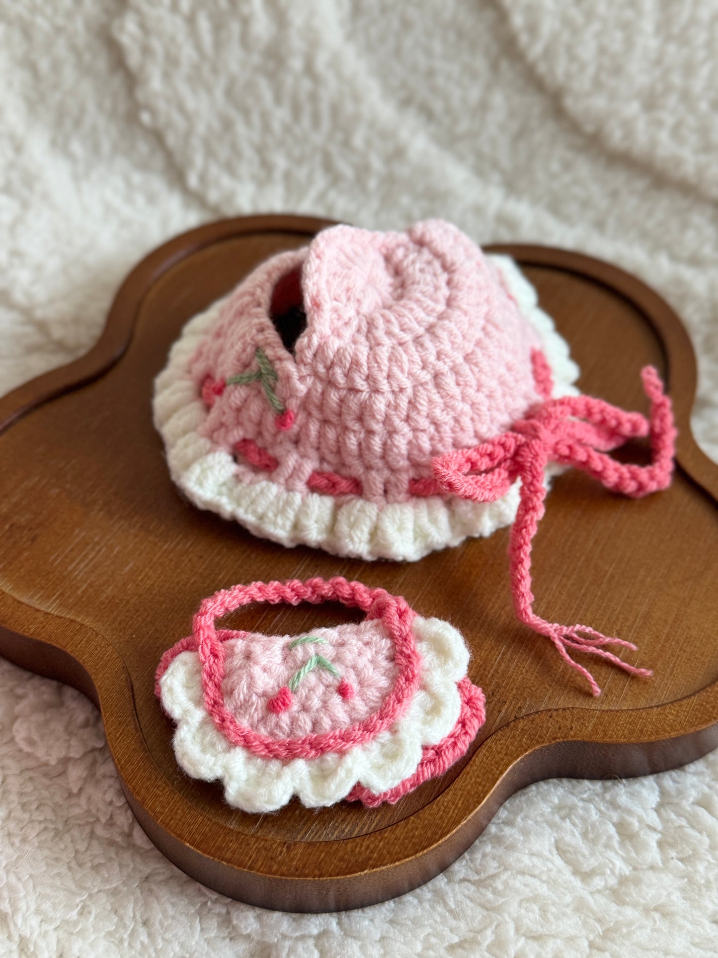 Flat lay of the cherry crochet bonnet and bib set displayed on a wooden flower-shaped tray.
