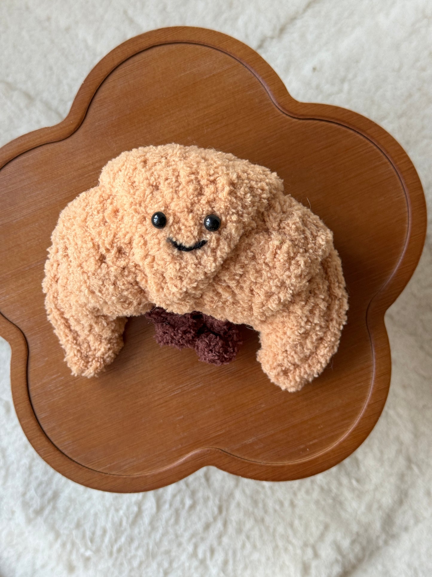 A handmade crochet version of the Jellycat croissant plush, featuring a textured tan body, smiling face with black eyes, and dark brown filling detail. Displayed on a wooden flower-shaped table with a soft cream background.