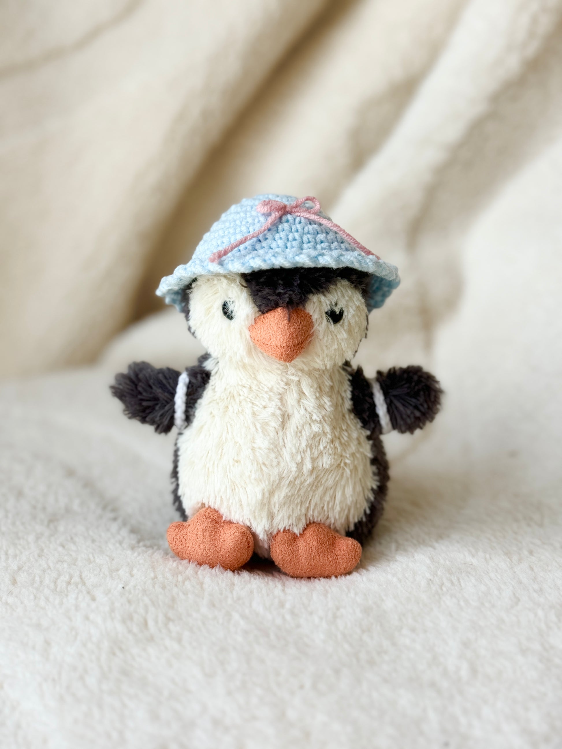 15 cm penguin plush wearing a sky-blue crocheted sun hat with a pink bow, seated on a cream blanket