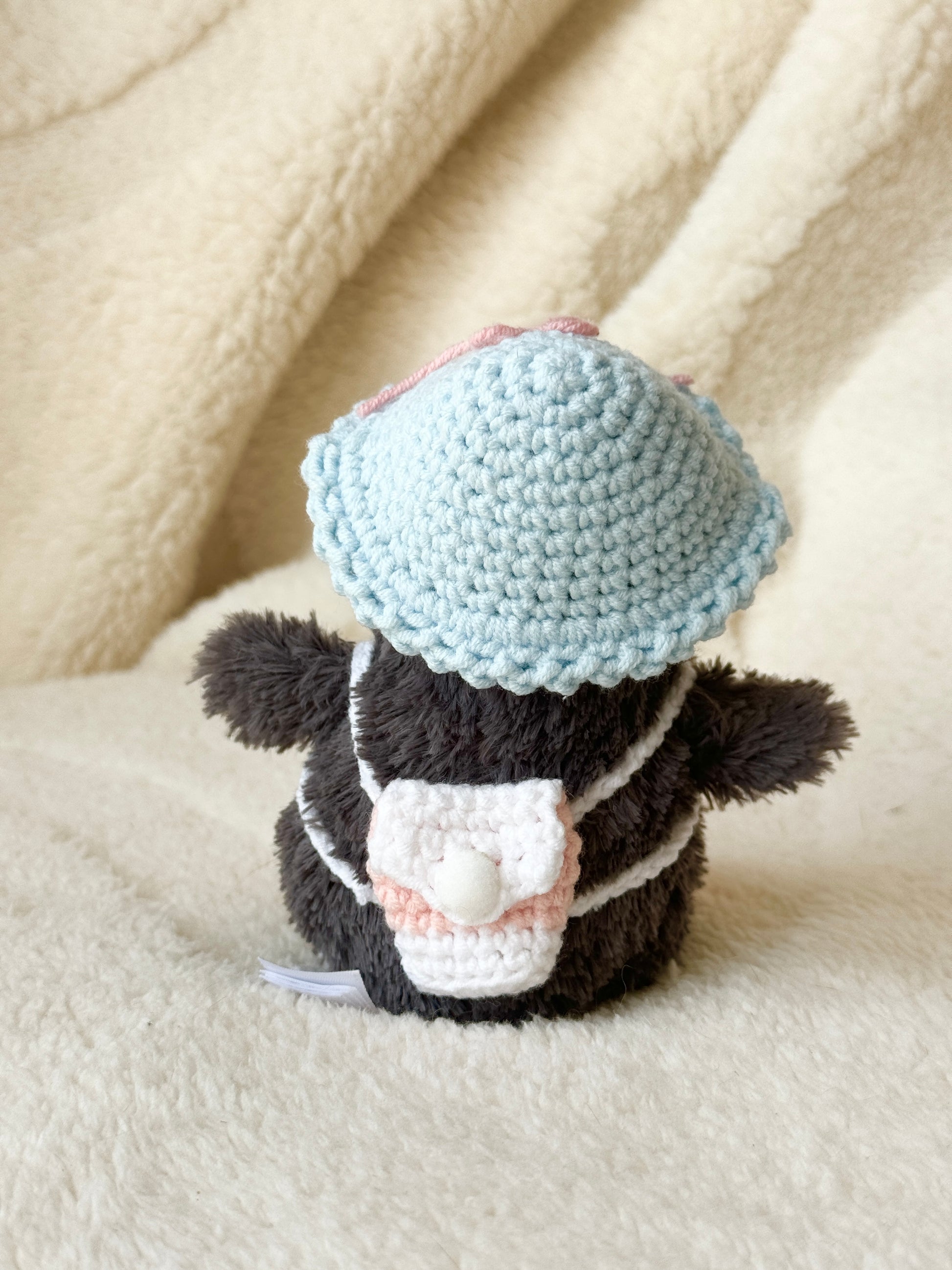Back of the penguin showing the tiny blush-and-cream crocheted backpack and blue hat on a cream blanket.
