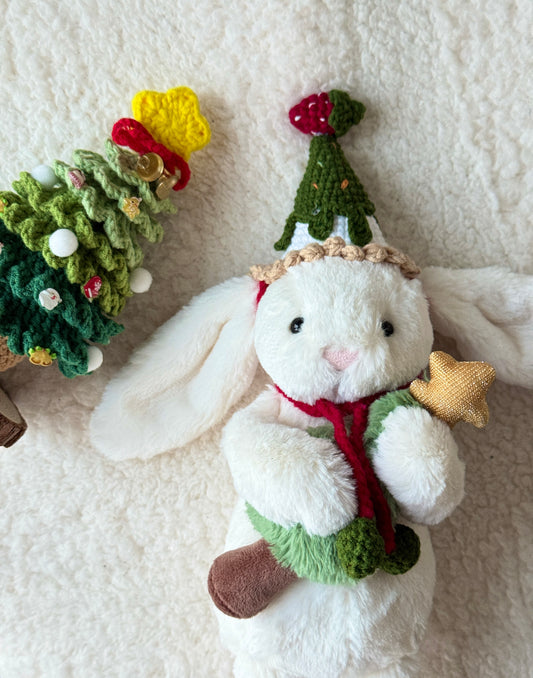 White Jellycat bunny wears a green crochet Christmas Tree hat with a cream ruffle brim and red ties; holding a plush tree with a gold star. A larger crochet tree decor sits nearby.