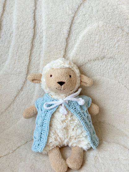 Jellycat Lollie Lamb (Small) wearing a handmade blue knit vest with white bow, seated on a fluffy cream background