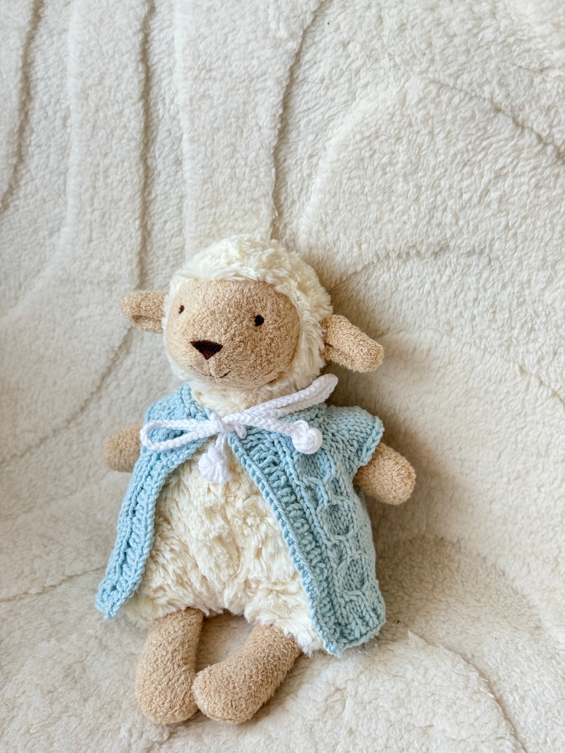 Side view of Jellycat Lollie Lamb dressed in a pale blue crochet vest with cable knit detail, shown on a cozy textured surface