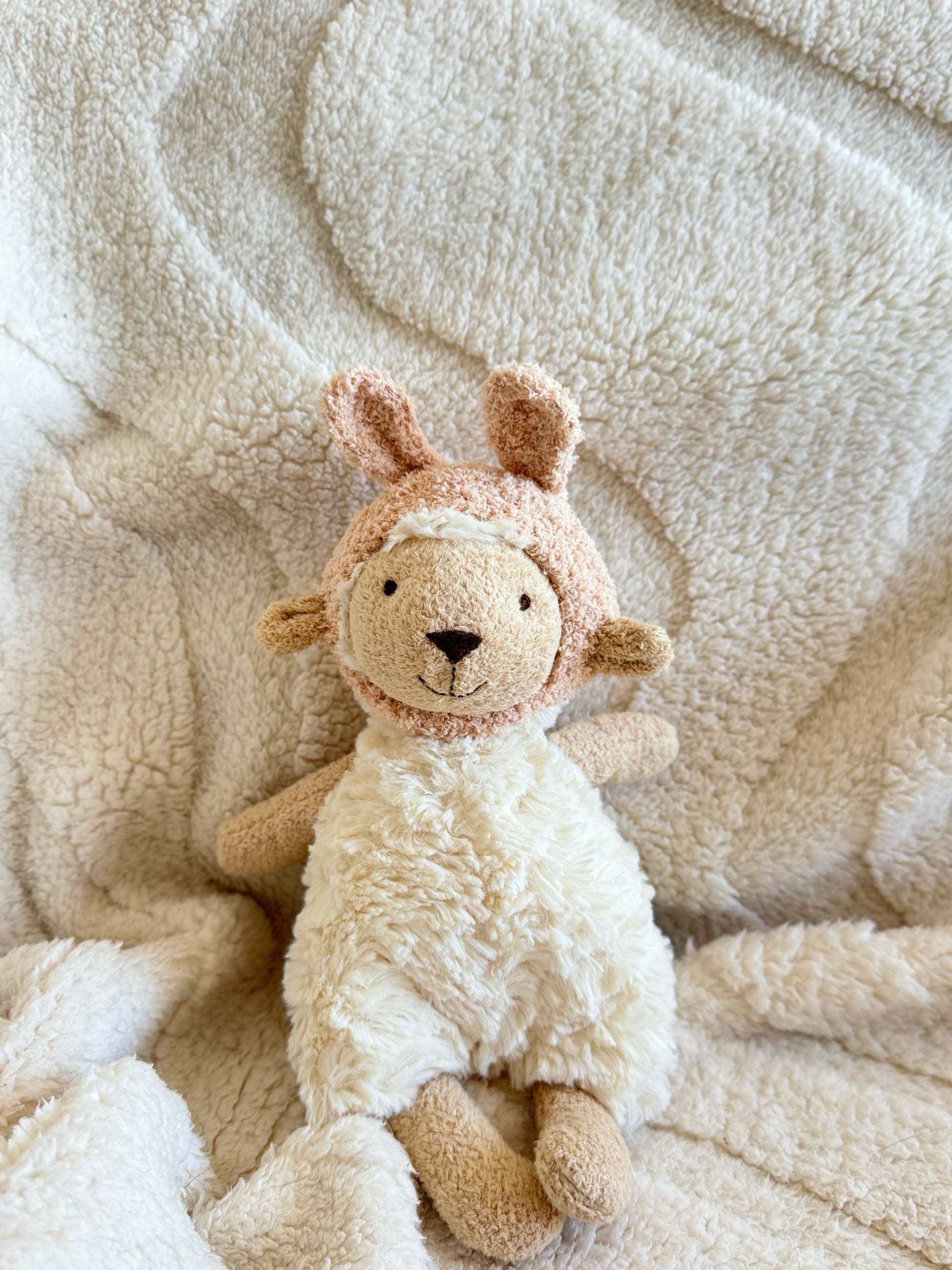 Jellycat Lottie Lamb wearing a soft pink bunny-ear hat, sitting on a cream blanket.