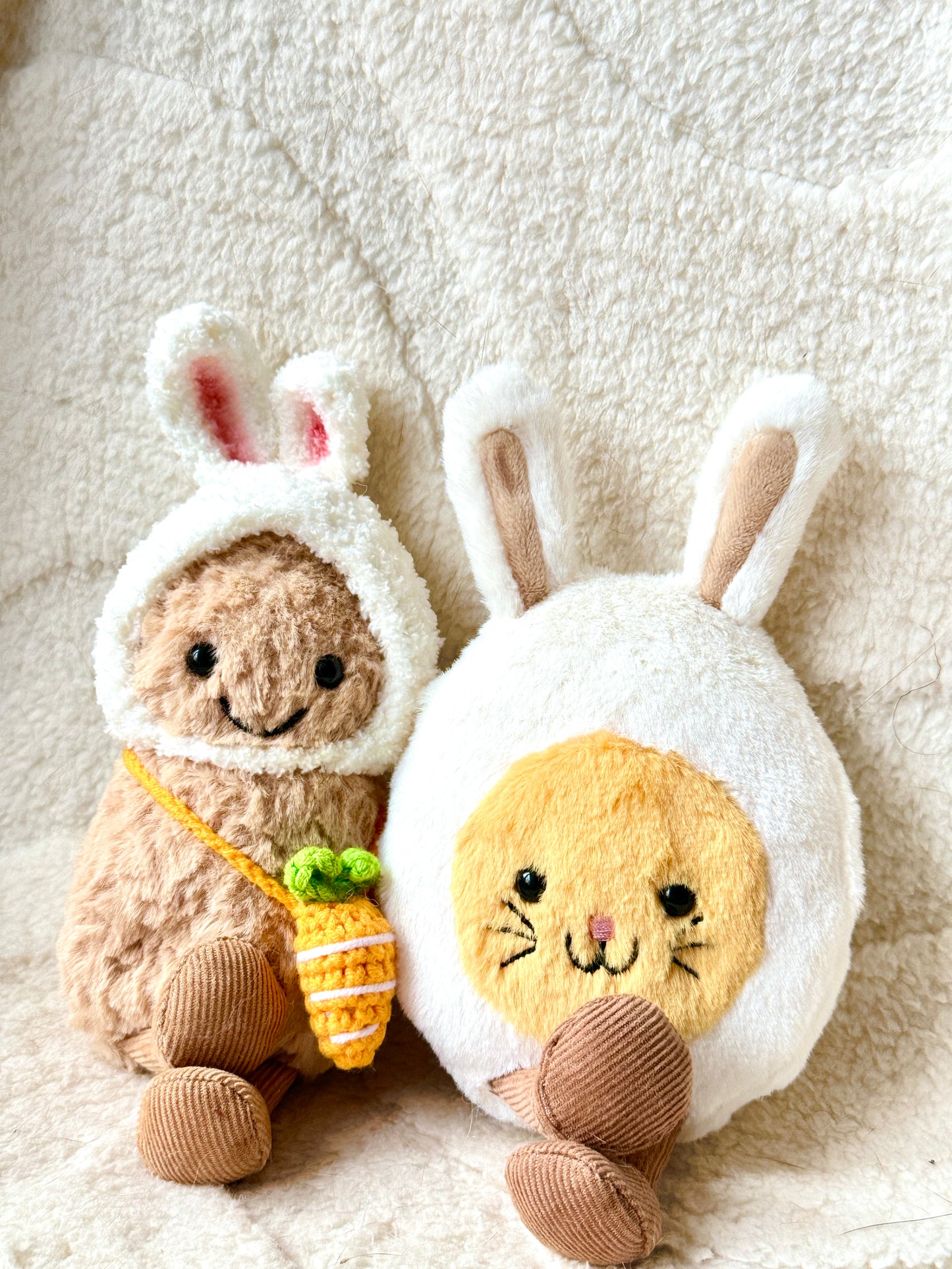 A Jellycat Little Peanut plush wearing a handmade fluffy white bunny hat with pink-lined ears and a small crochet carrot bag slung across its body. The plush sits next to a Jellycat bunny egg plush, both placed on a soft, neutral-toned fabric background with delicate white daisy ribbon accents. 
