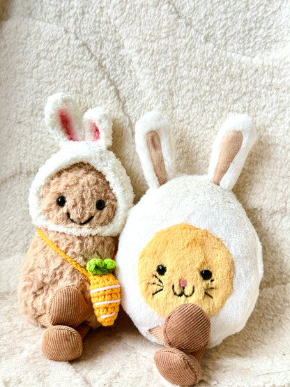 A Jellycat Little Peanut plush wearing a handmade fluffy white bunny hat with pink-lined ears and a small crochet carrot bag slung across its body. The plush sits next to a Jellycat bunny egg plush, both placed on a soft, neutral-toned fabric background with delicate white daisy ribbon accents. 