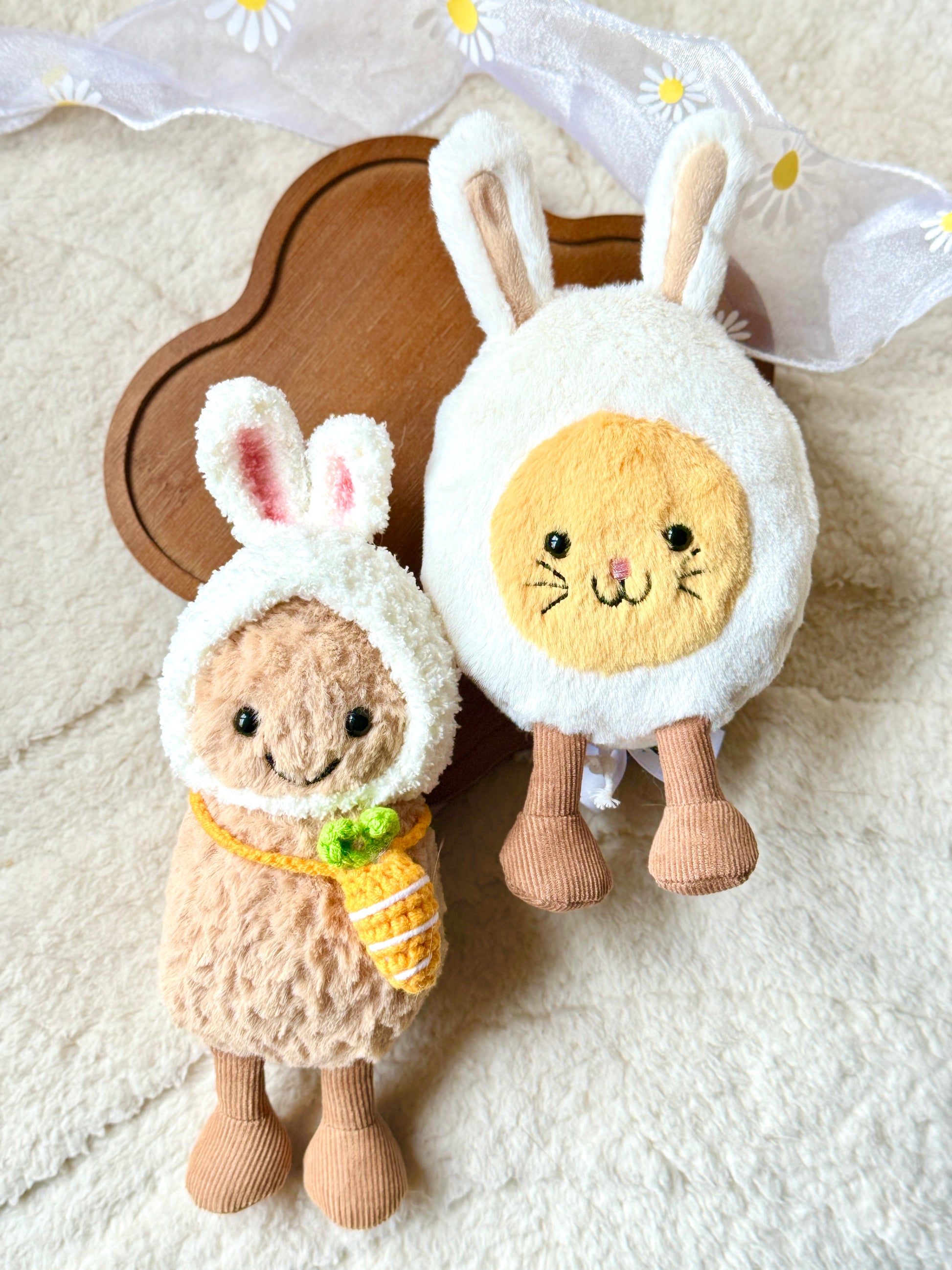 A Jellycat Little Peanut plush wearing a handmade fluffy white bunny hat with pink-lined ears and a small crochet carrot bag slung across its body. The plush sits next to a Jellycat bunny egg plush, both placed on a soft, neutral-toned fabric background with delicate white daisy ribbon accents. 