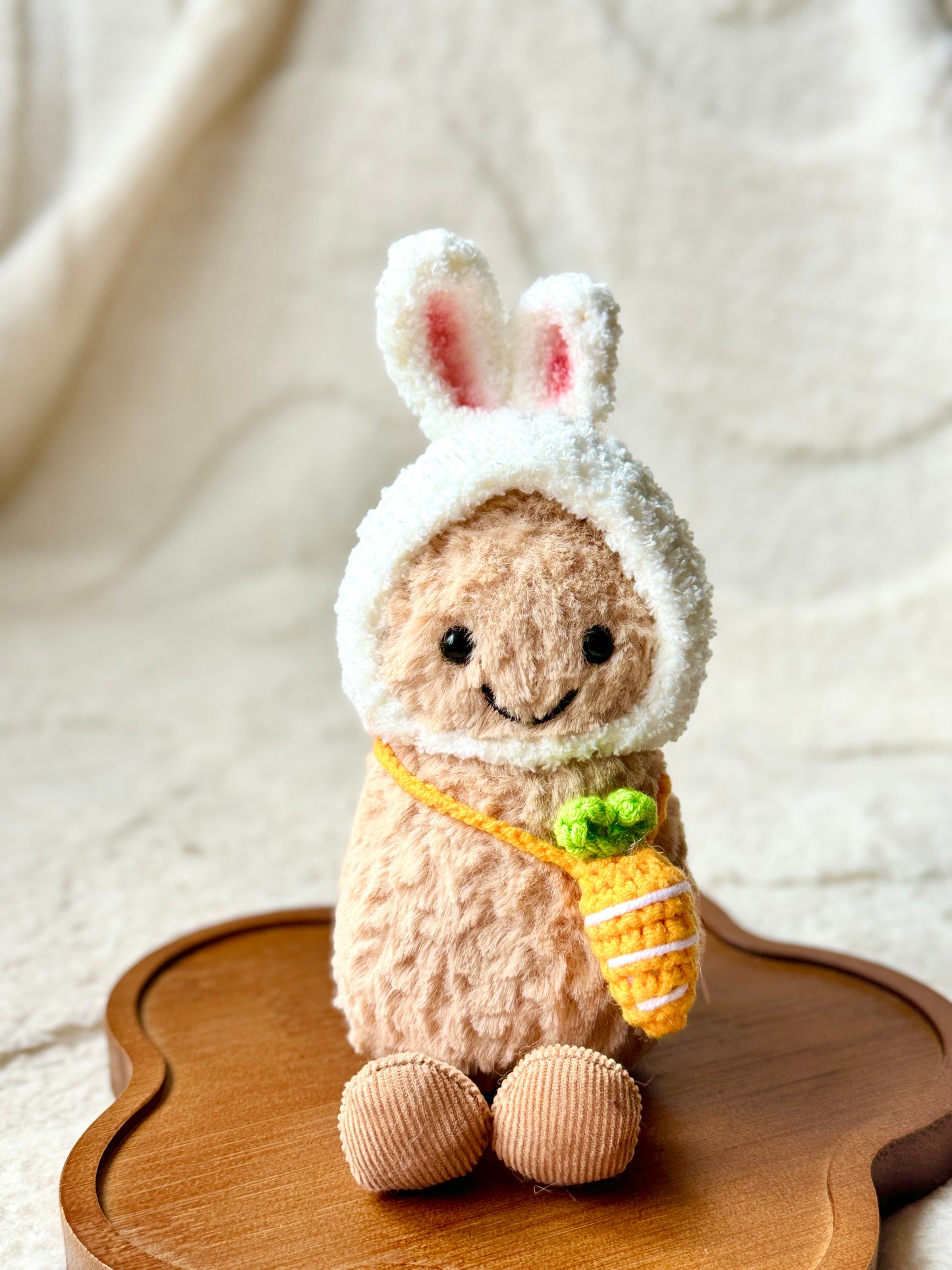 A Jellycat Little Peanut plush wearing a handmade fluffy white bunny hat with pink-lined ears and a small crochet carrot bag slung across its body. 