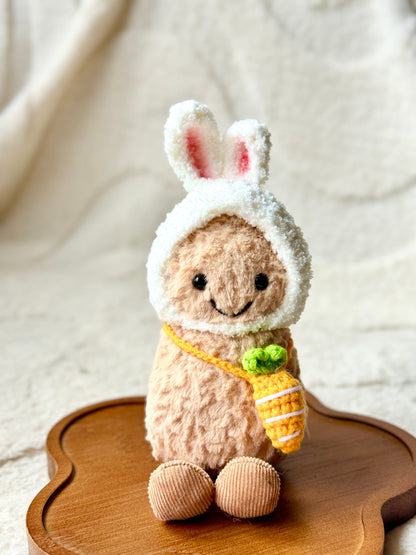 A Jellycat Little Peanut plush wearing a handmade fluffy white bunny hat with pink-lined ears and a small crochet carrot bag slung across its body. 