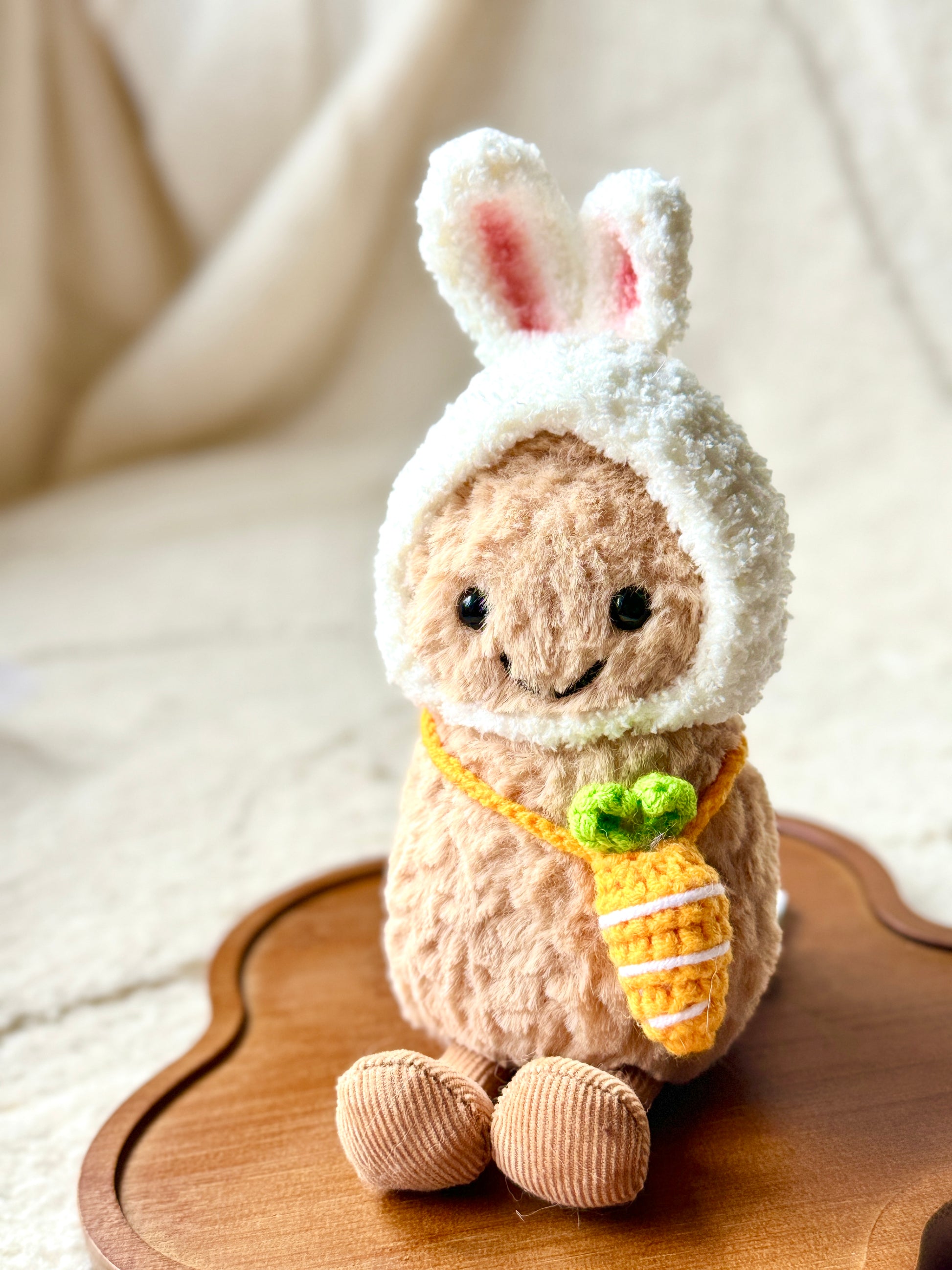 A Jellycat Little Peanut plush wearing a handmade fluffy white bunny hat with pink-lined ears and a small crochet carrot bag slung across its body. T
