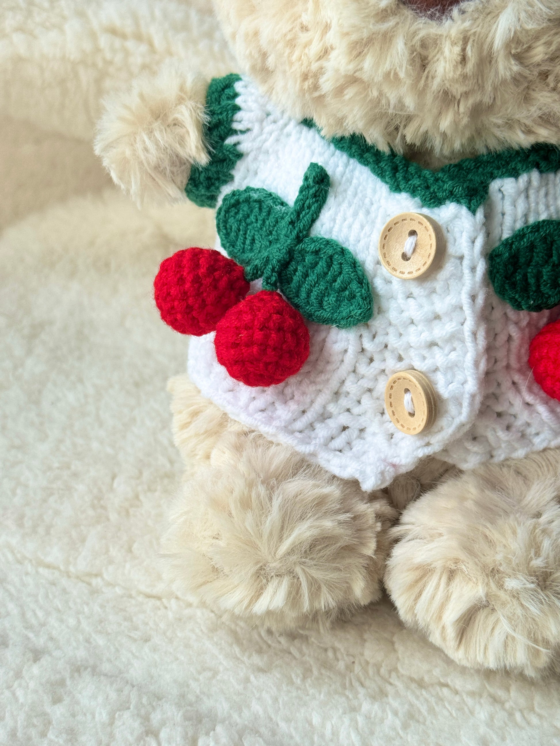 Close-up of a handmade cherry cardigan for Jellycat Bart Junior, showing white crochet texture, green trim, wooden-style buttons, and red crochet cherry details.