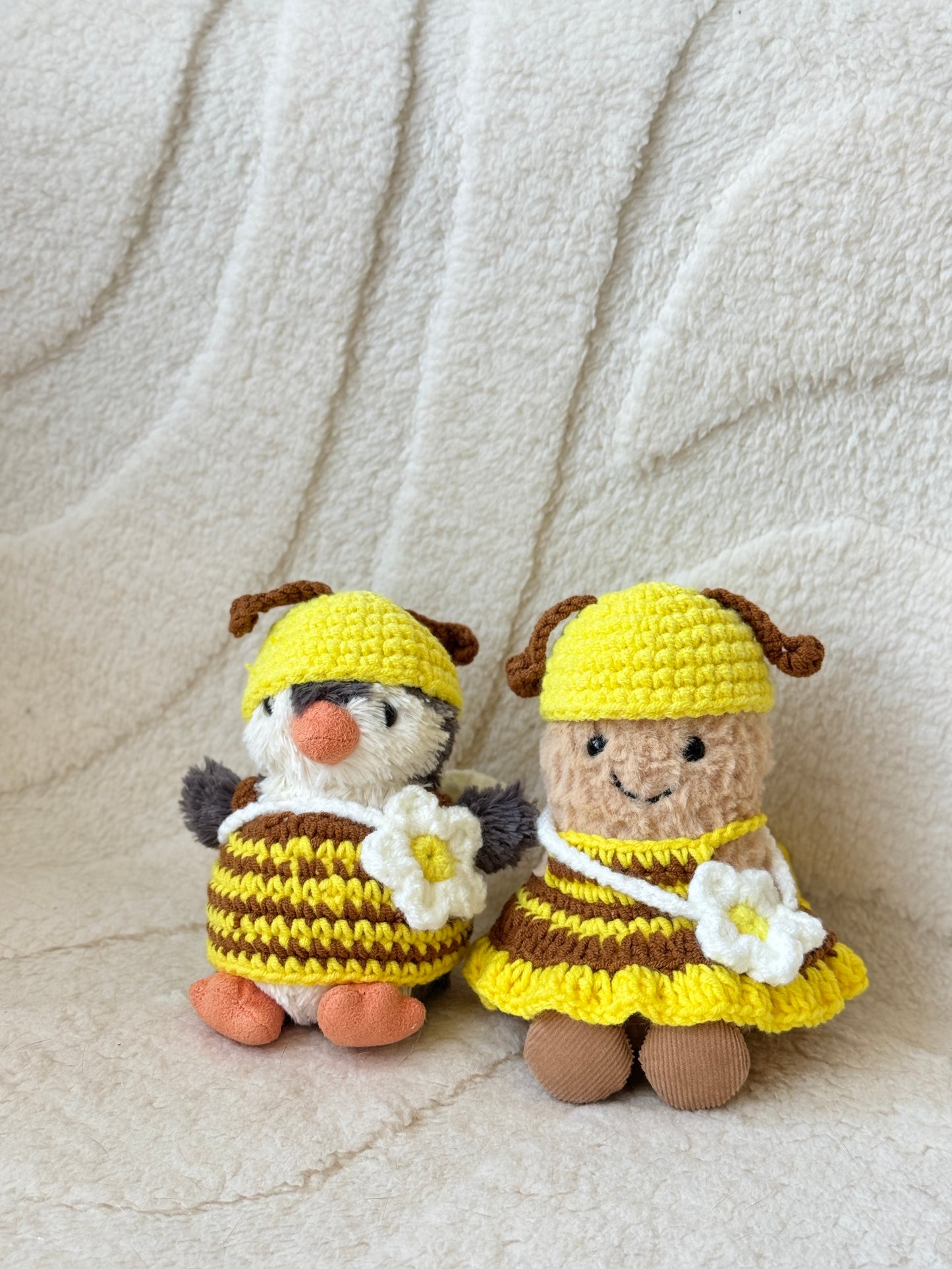 Two Jellycat plush toys—Peanut and Penguin—dressed in matching handmade crochet bee outfits with yellow and brown striped dresses, yellow hats with brown antennae, and white flower crossbody bags. They are sitting side by side on a soft cream background.