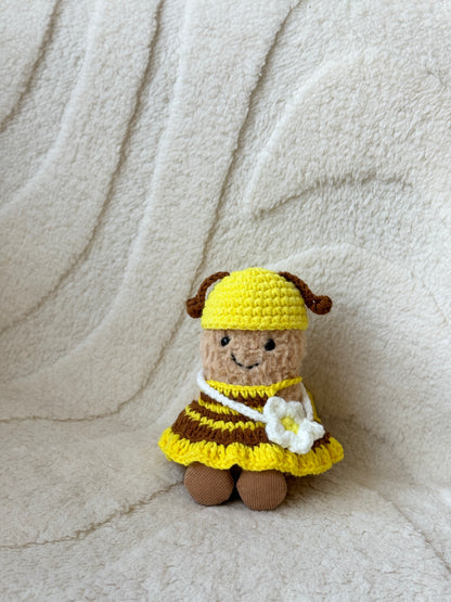 Jellycat Peanut plush dressed in a handmade crochet bee outfit, featuring a yellow and brown striped dress, a yellow hat with brown antennae, and a white flower crossbody bag. Posed solo against a cream textured fabric background.

