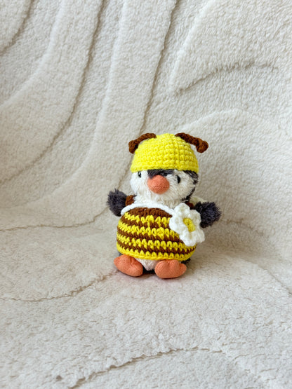 Jellycat Penguin plush dressed in a handmade crochet bee outfit, featuring a yellow and brown striped dress, a yellow hat with brown antennae, and a white flower crossbody bag. Posed solo against a cream textured fabric background.

