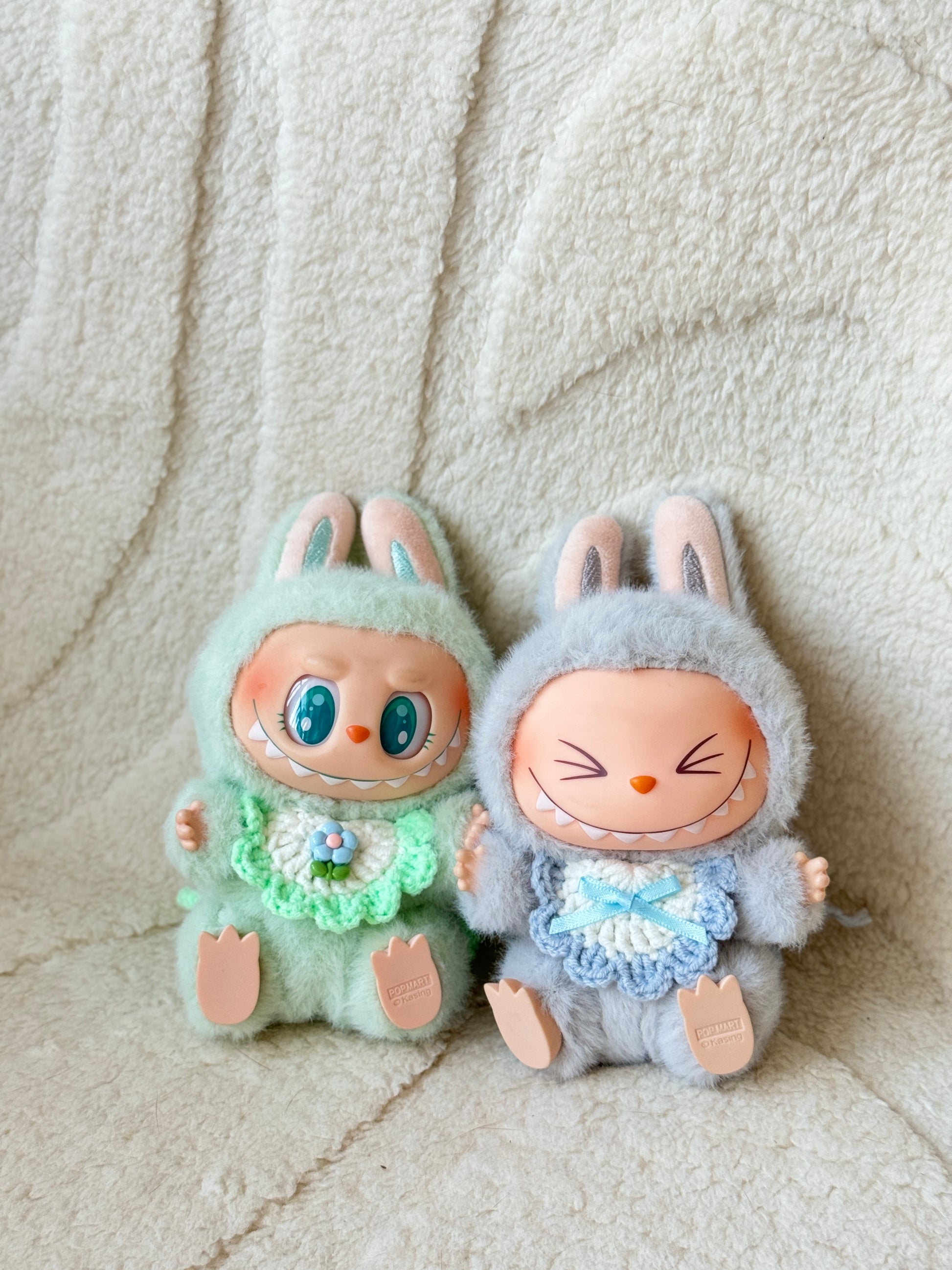 Two Pop Mart Labubu plush figures wearing handmade crochet bibs—one in mint green with a flower, the other in pastel blue with a bow—posed on a soft cream background.