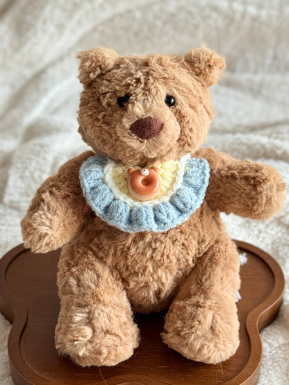 Jellycat Bartholomew Bear wearing a pastel crochet bib with a ceramic donut bear charm, seated on a wooden tray.
