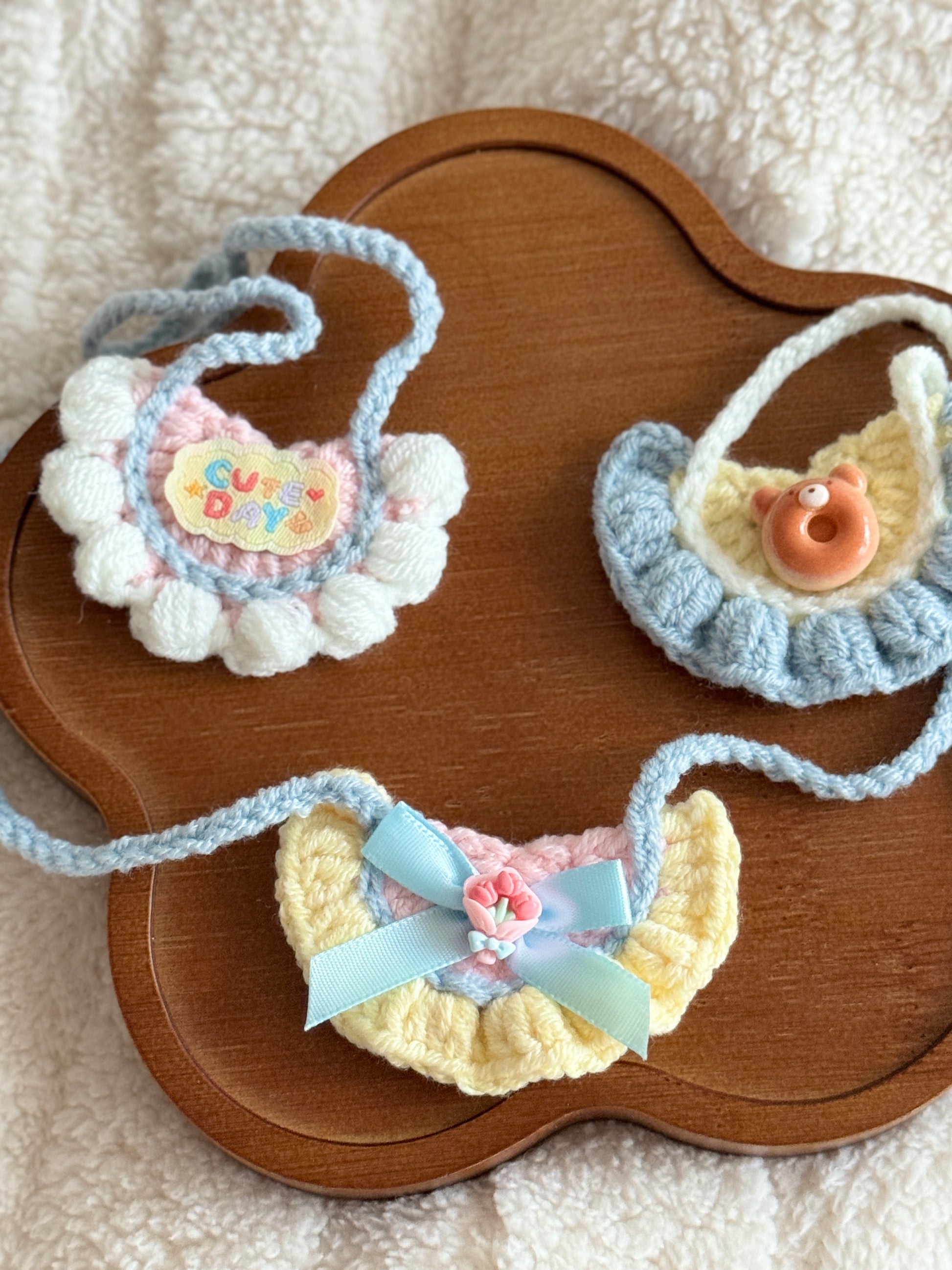 Three handmade crochet bibs displayed on a wooden tray, showing designs with a bear charm, “Cute Day” label, and bow.

