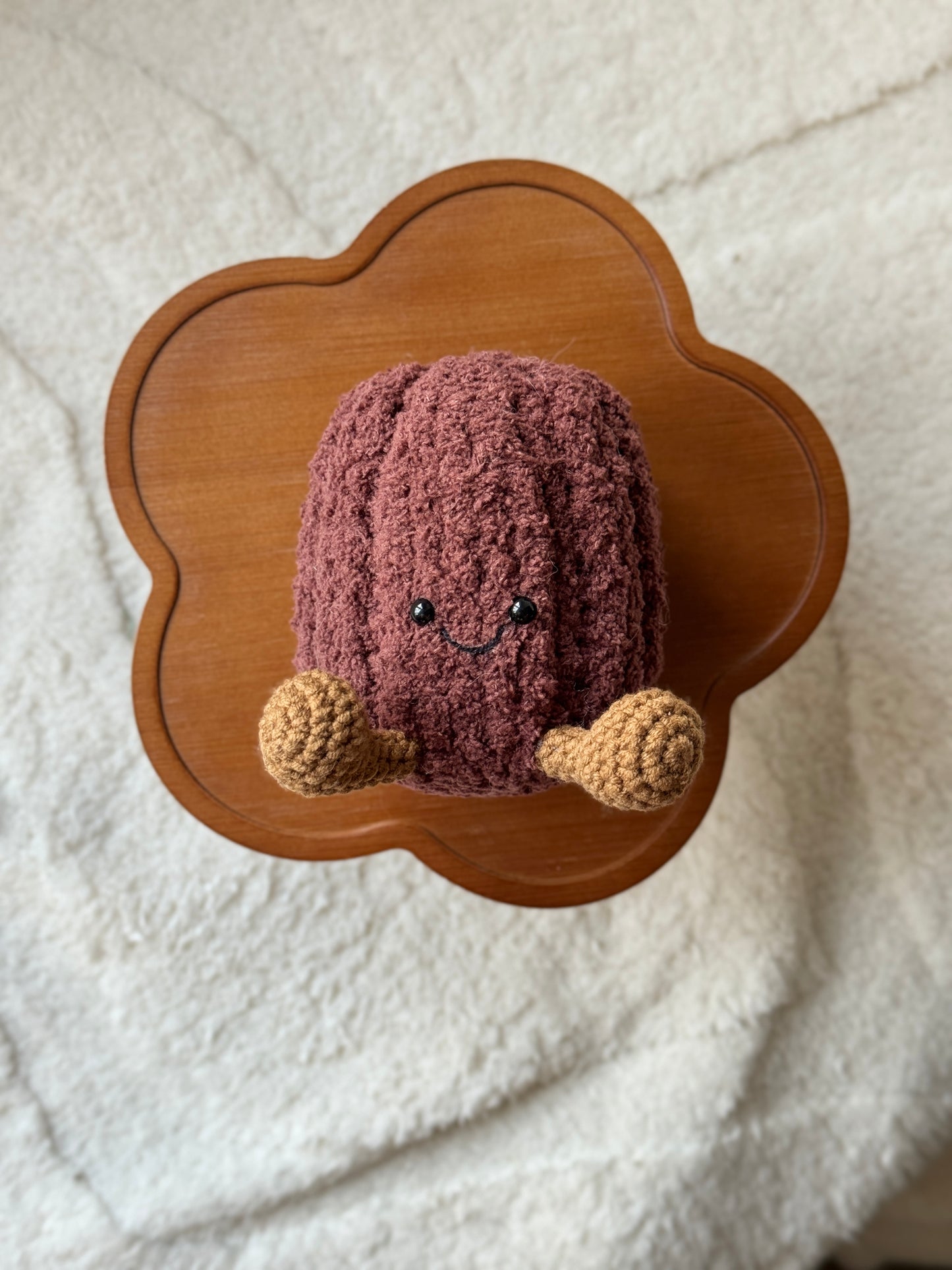 Crochet canelé plushie with a ribbed chocolate-brown body, smiley face, and tan legs, sitting on a wooden clover-shaped stand.