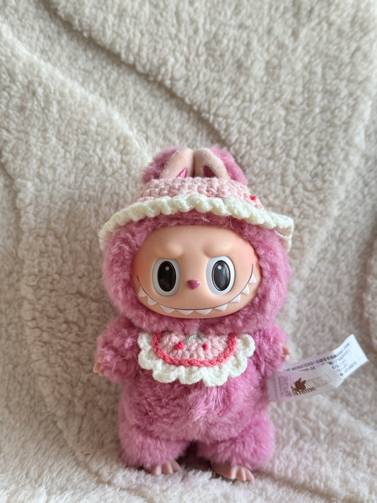 Labubu Monster Meimei wearing a handmade cherry-themed bonnet and bib crochet outfit, standing on a soft cream blanket.