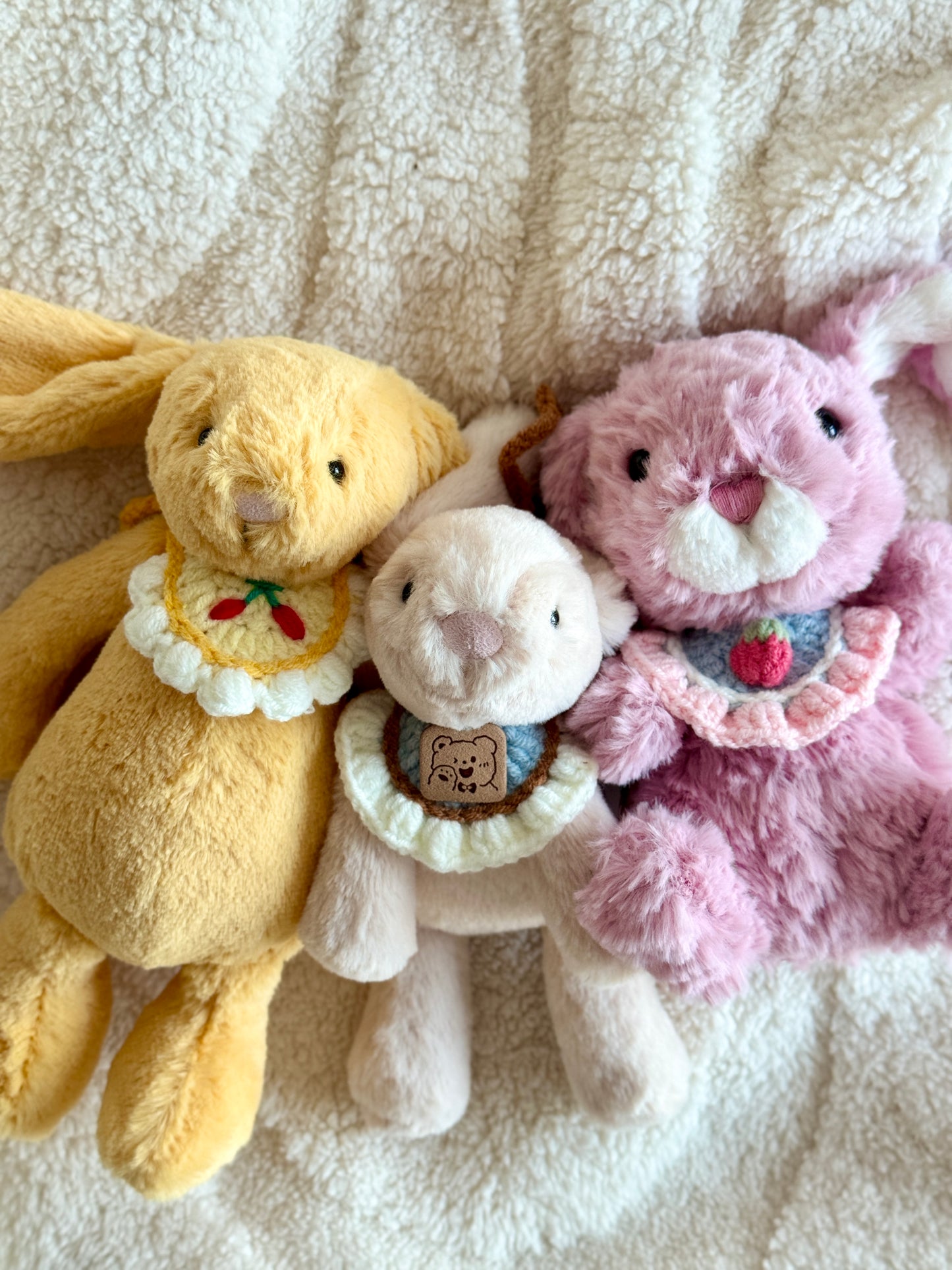 Three Jellycat plush bunnies wearing handmade crochet collars, sitting close together on a cream sherpa blanket. The collars feature a cherry motif, a strawberry motif, and a bear face patch.