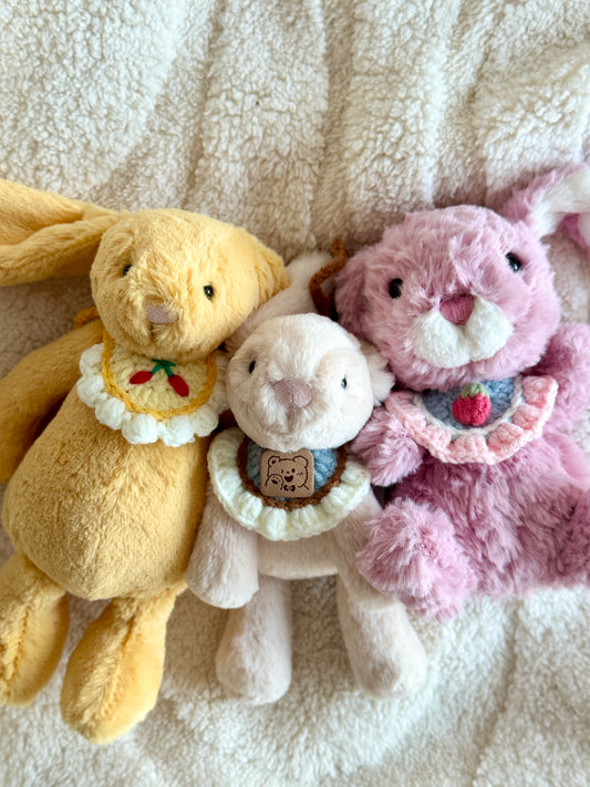 Three Jellycat plush bunnies wearing handmade crochet collars, sitting close together on a cream sherpa blanket. The collars feature a cherry motif, a strawberry motif, and a bear face patch.