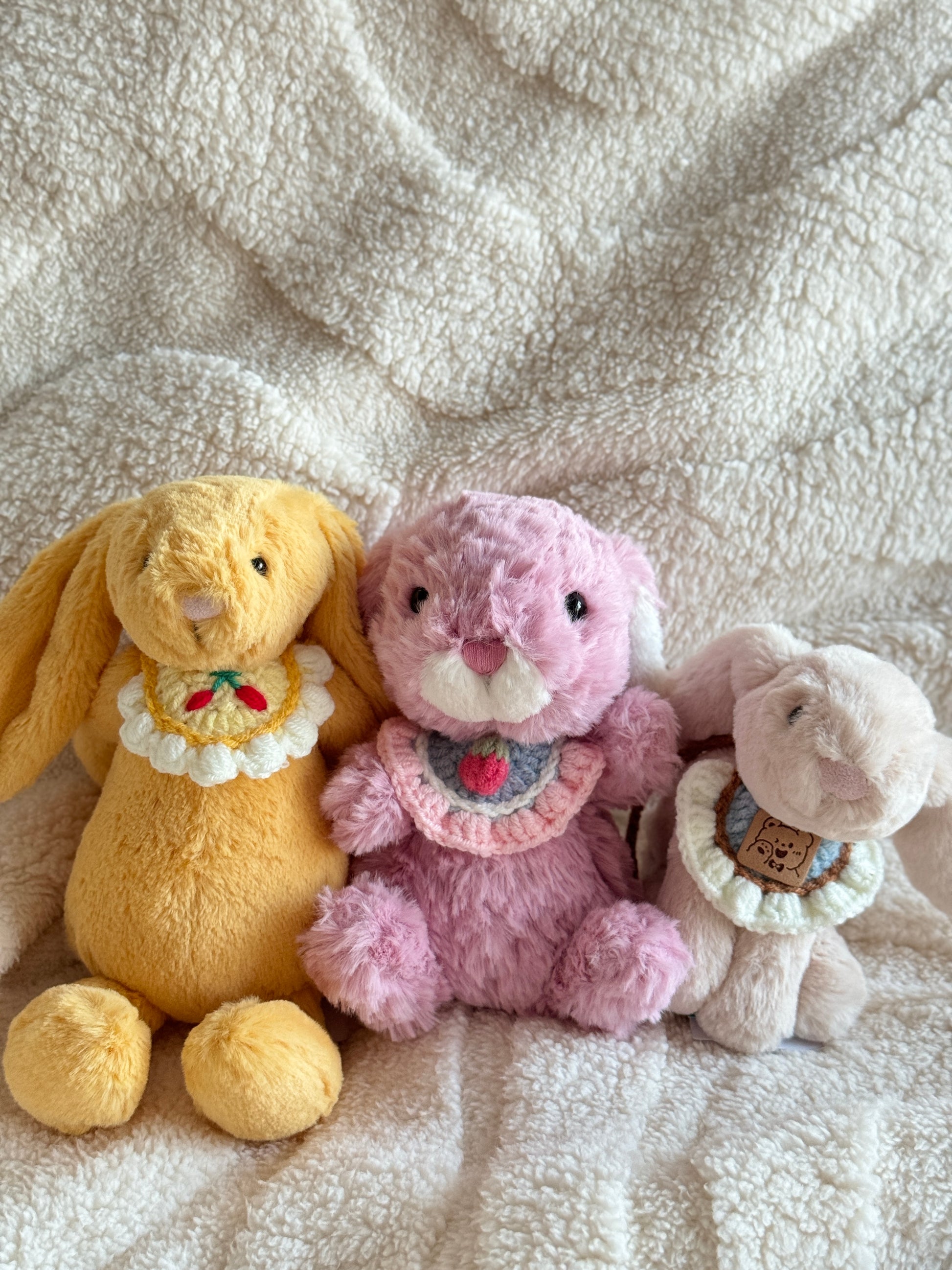 Jellycat bunnies in yellow, pink, and cream lined up side by side on a fluffy white blanket, each wearing a unique crochet bib with fruit or bear designs.