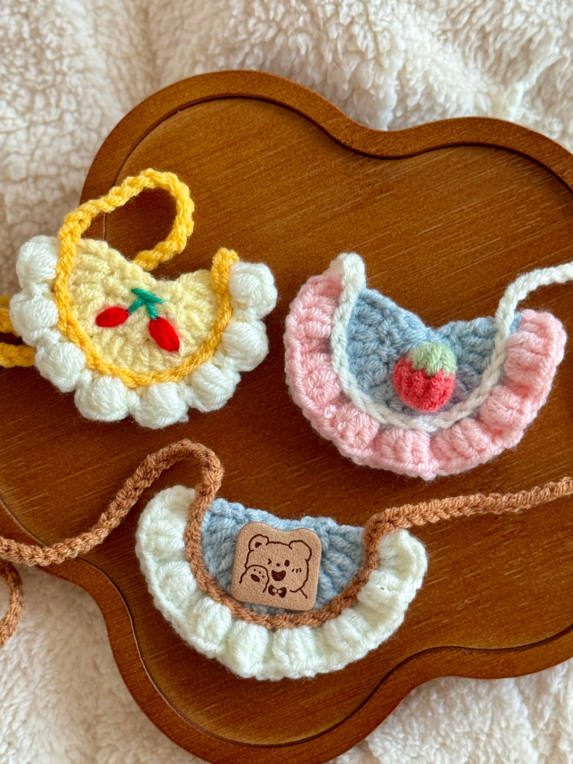 Flat lay of three handmade crochet collars for Jellycat plushies, displayed on a wooden tray. Designs include cherries, a strawberry, and a leather bear patch.
