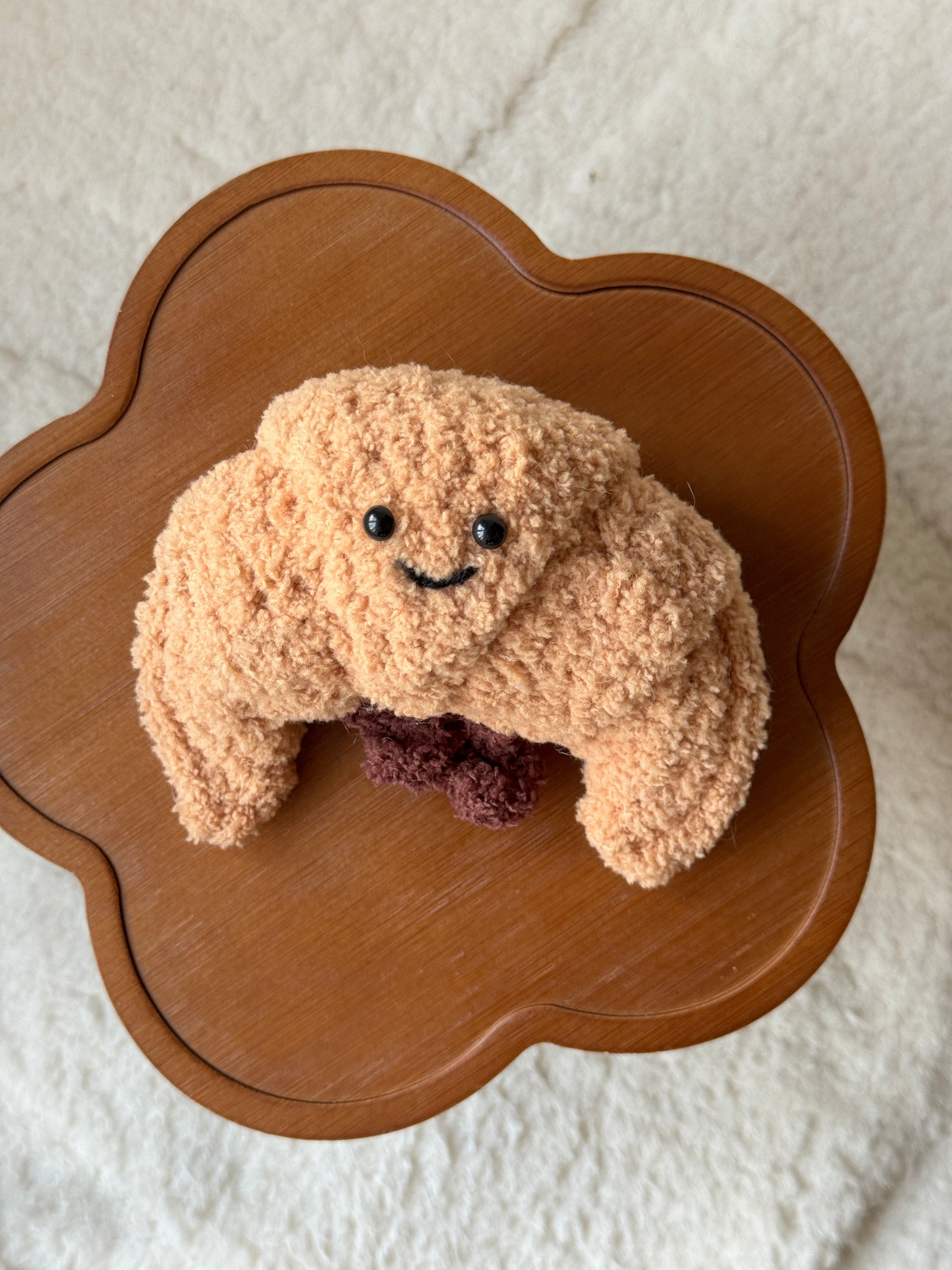 A handmade crochet version of the Jellycat croissant plush, featuring a textured tan body, smiling face with black eyes, and dark brown filling detail. Displayed on a wooden flower-shaped table with a soft cream background.
