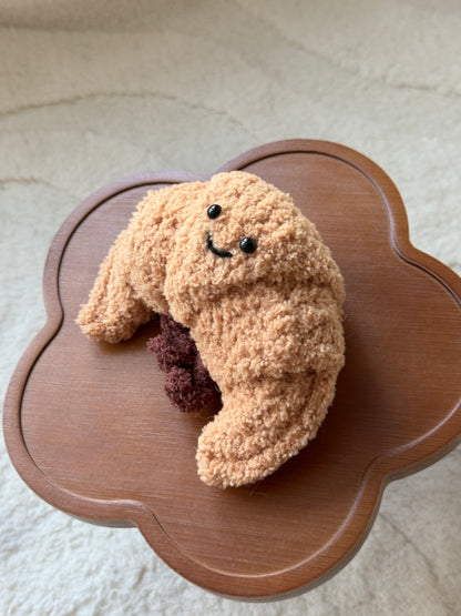 A handmade crochet version of the Jellycat croissant plush, featuring a textured tan body, smiling face with black eyes, and dark brown filling detail. Displayed on a wooden flower-shaped table with a soft cream background.