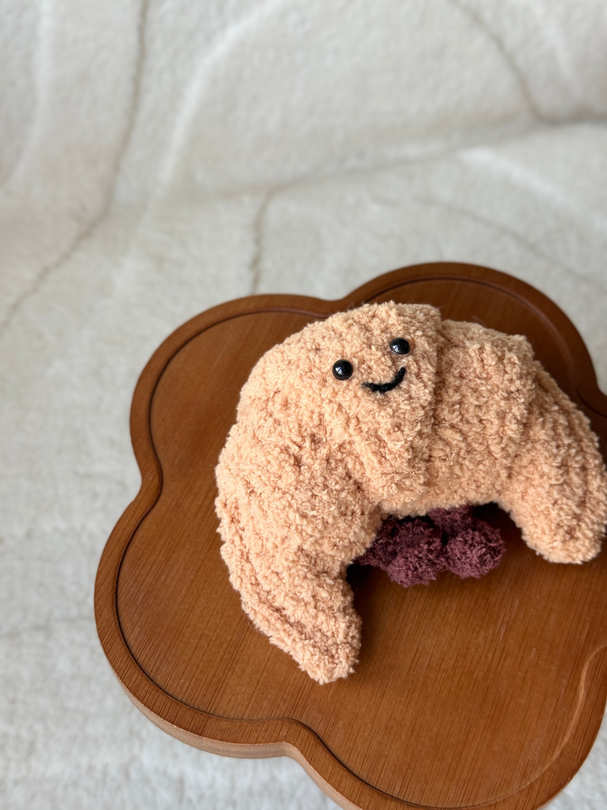 A handmade crochet version of the Jellycat croissant plush, featuring a textured tan body, smiling face with black eyes, and dark brown filling detail. Displayed on a wooden flower-shaped table with a soft cream background.