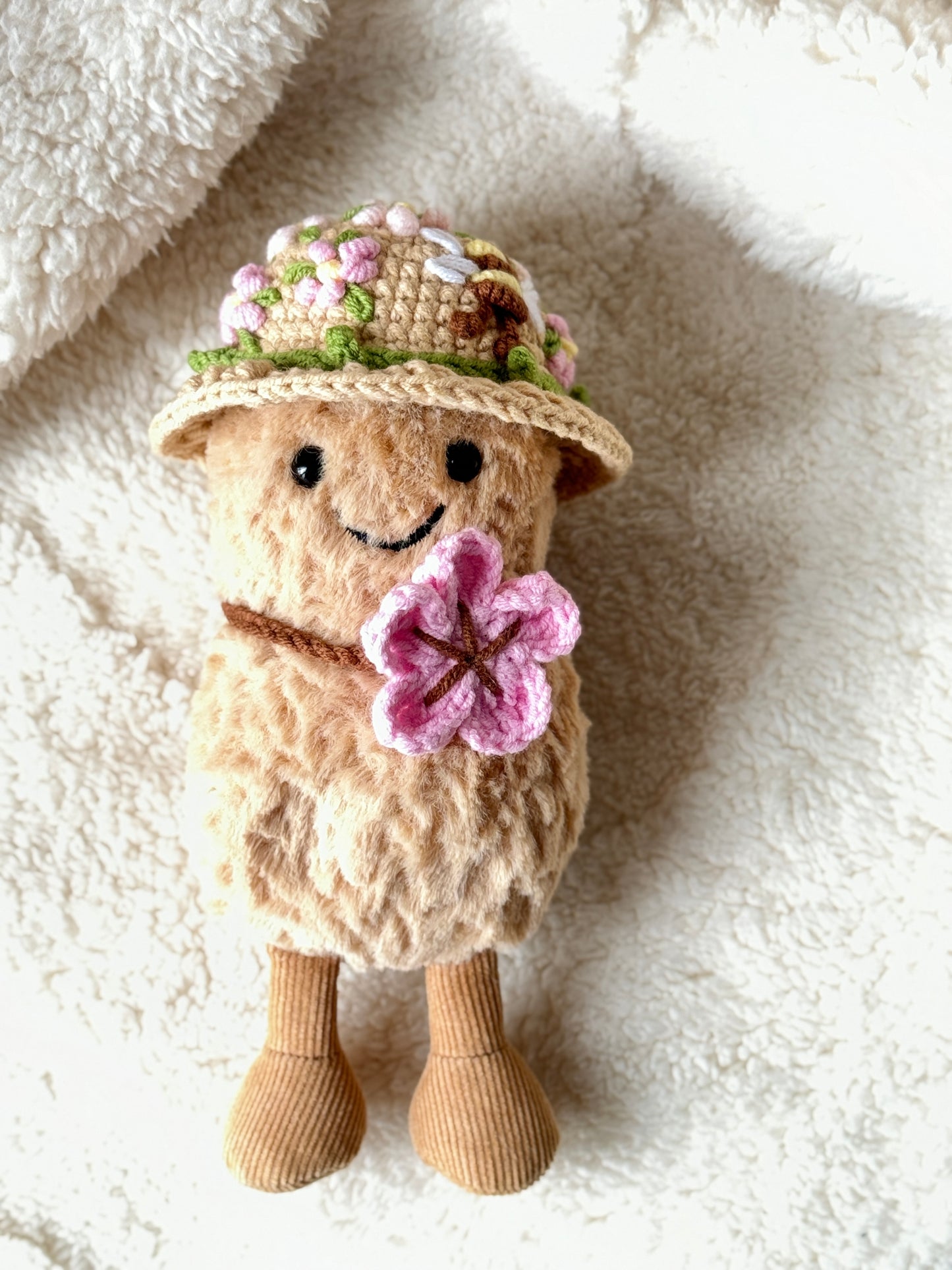 Jellycat Amuseable Peanut wearing a handmade crochet floral hat with blossoms and a bee, holding a pink crochet flower.