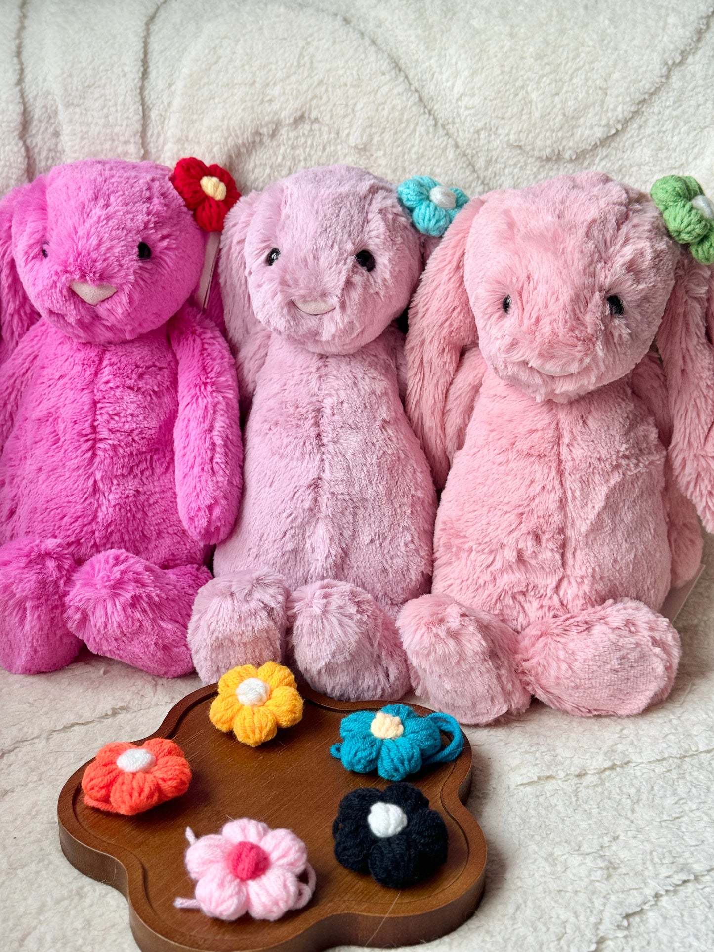 Three Jellycat Bashful Bunnies sitting upright, each adorned with a different crochet flower accessory. A wooden tray in front of them holds additional crochet flowers, displaying the variety of colors available.