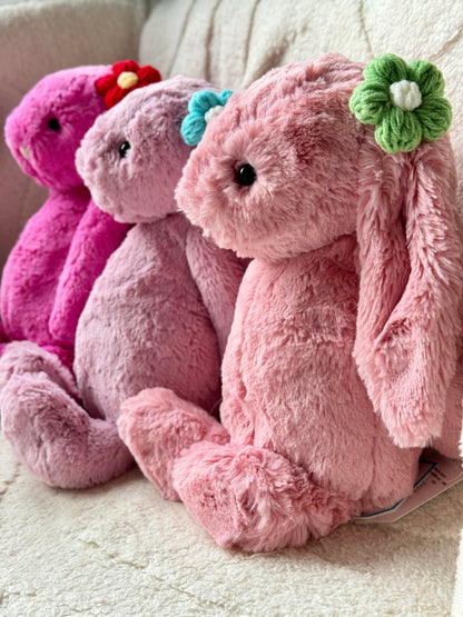 Three Jellycat Bashful Bunnies in shades of pink, sitting in a row on a soft cream-colored surface. Each bunny wears a handmade crochet flower hair accessory in different colors—green, blue, and red.

