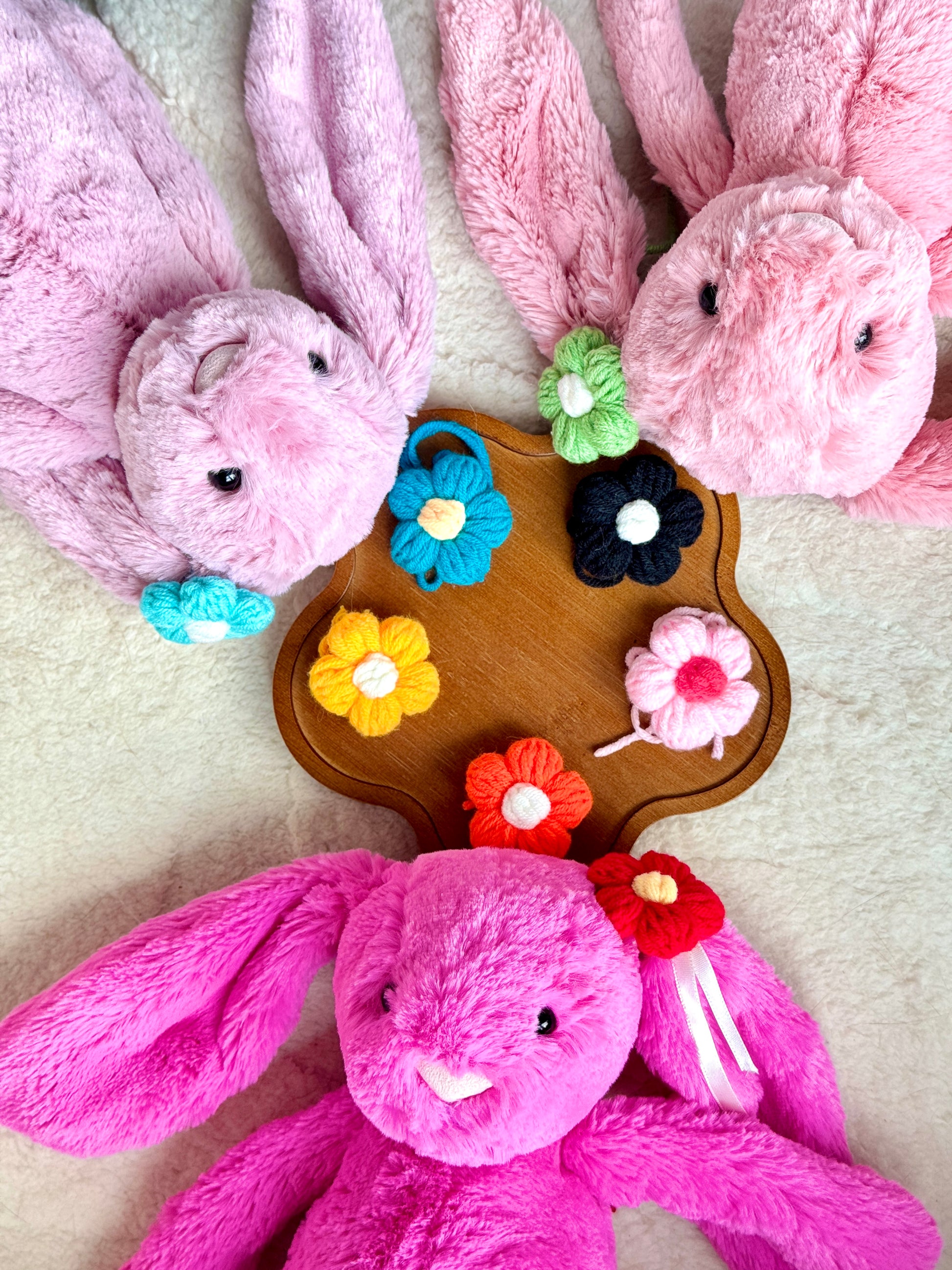 A top-down view of three Jellycat Bunnies lying on their backs with floppy ears spread out. A wooden tray holds an assortment of colorful crochet flower hair accessories, showcasing different designs and colors.