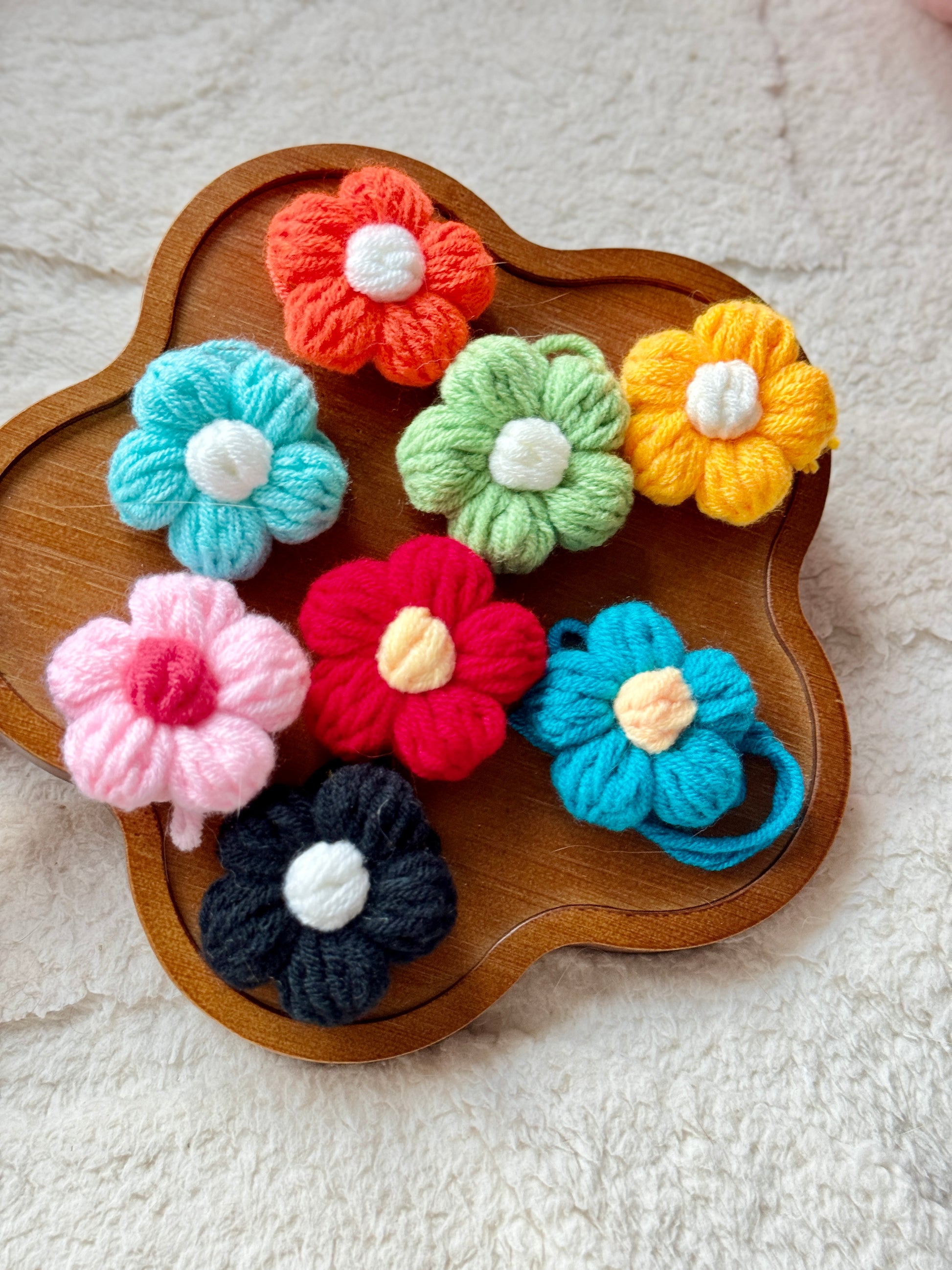 A close-up of a wooden tray displaying seven handmade crochet flower hair accessories in various colors, including red, green, yellow, pink, black, and blue. Each flower is crafted with soft yarn and features a small white center.
