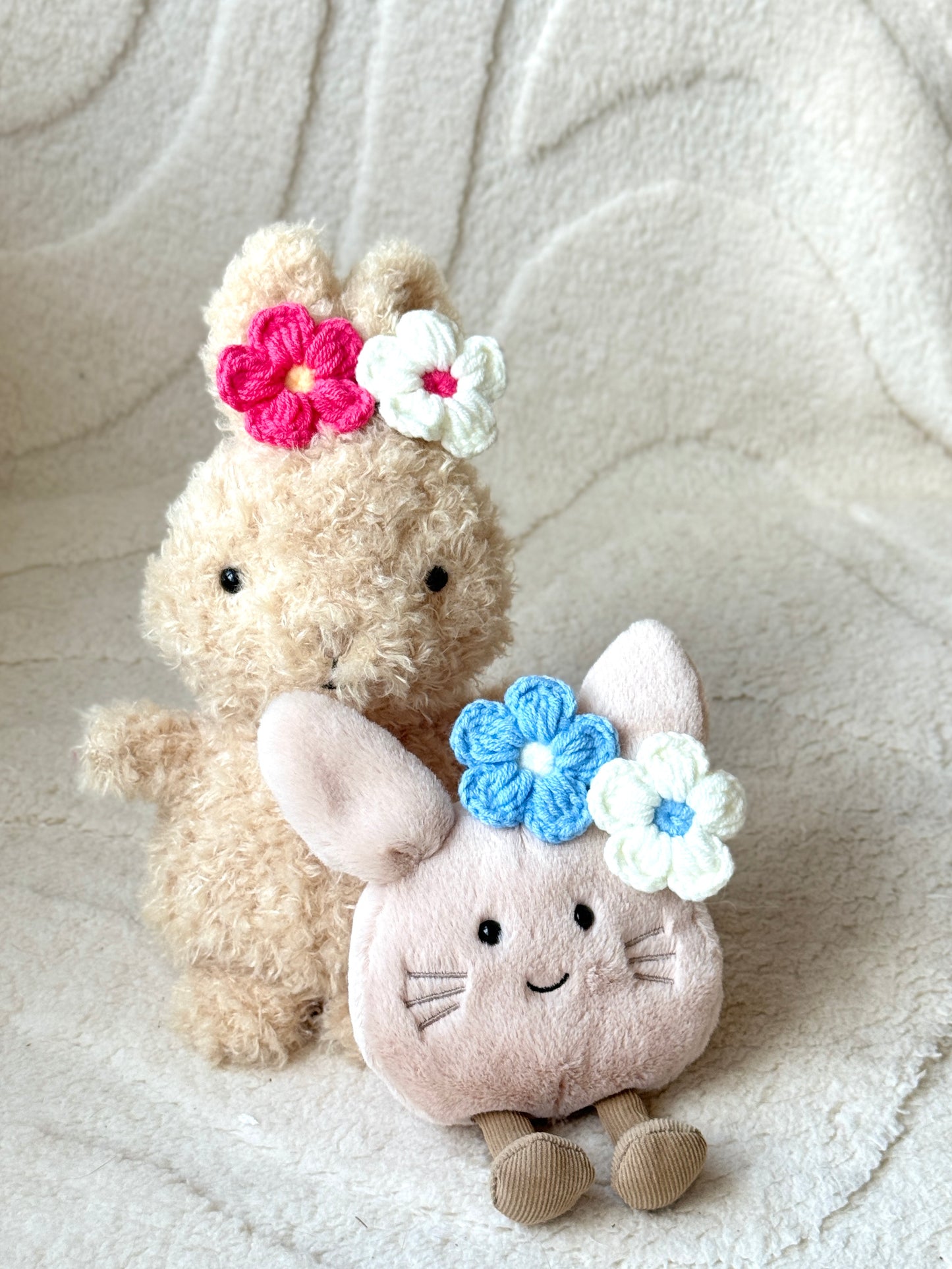 Two Jellycat plush toys wearing colorful crochet flower clips—pink, red, blue, and white—styled on their heads.
