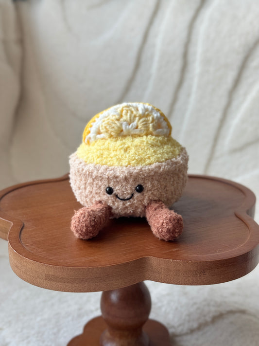 Handmade crochet lemon tart plush with a yellow top, brown legs, and a smiling face, sitting on a wooden clover-shaped stand.