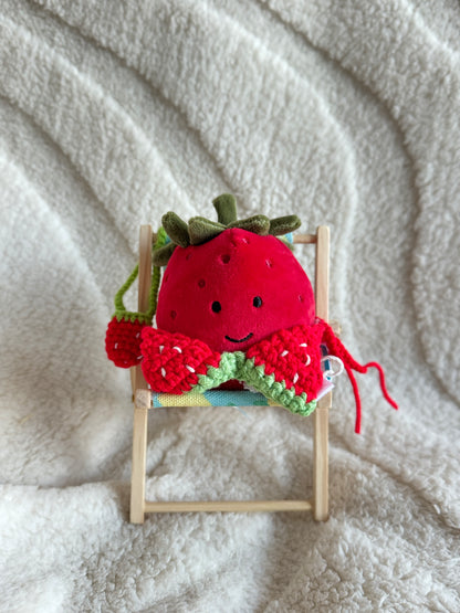 Jellycat strawberry plush wearing a red crochet bikini top with green trim, seated in a mini deck chair.