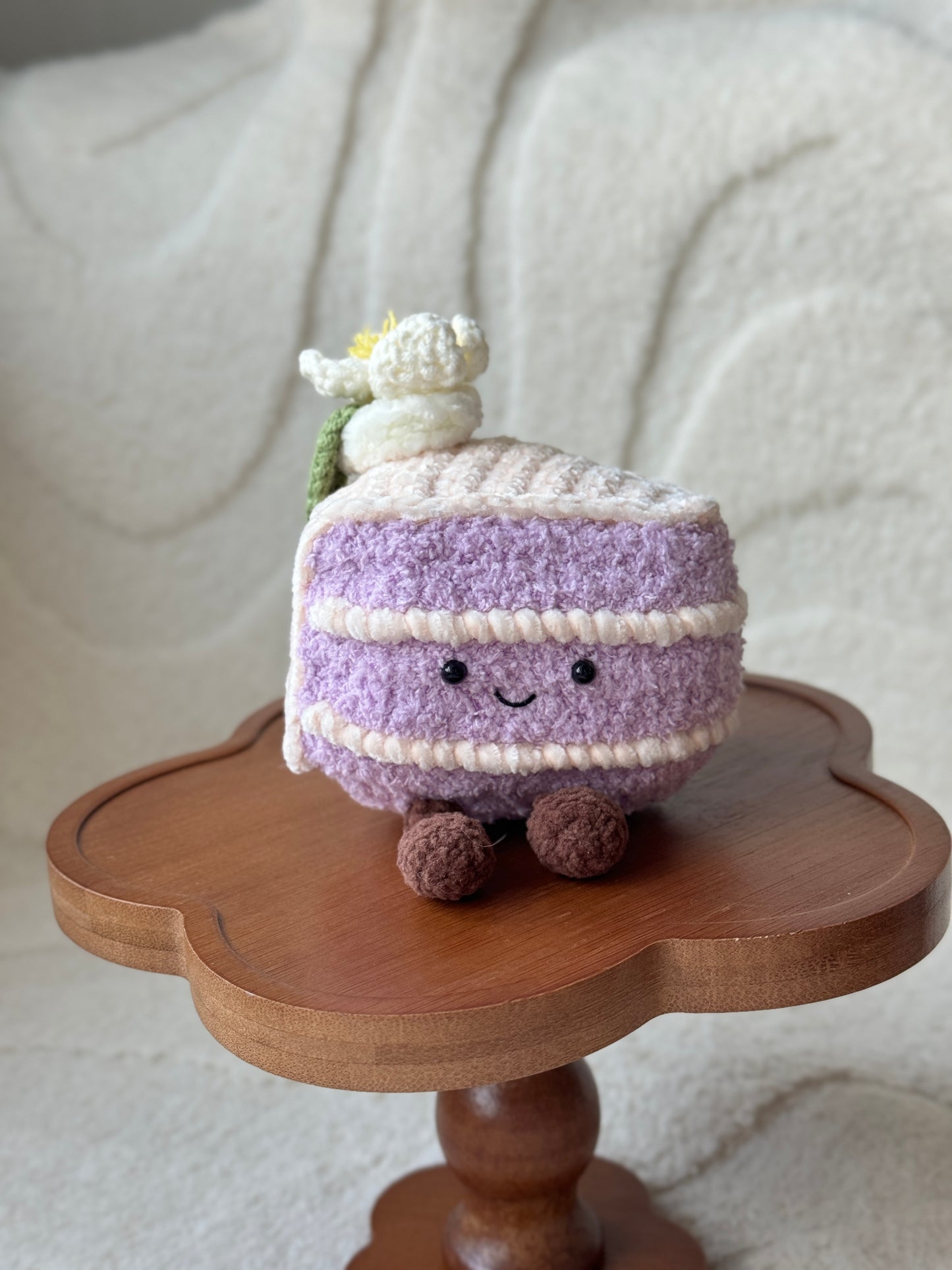 crochet taro cake plush inspired by Jellycat’s Shanghai-exclusive Amuseable Taro Cake. Features lavender layers, a white flower topper, and a smiling face, placed on a wooden flower-shaped