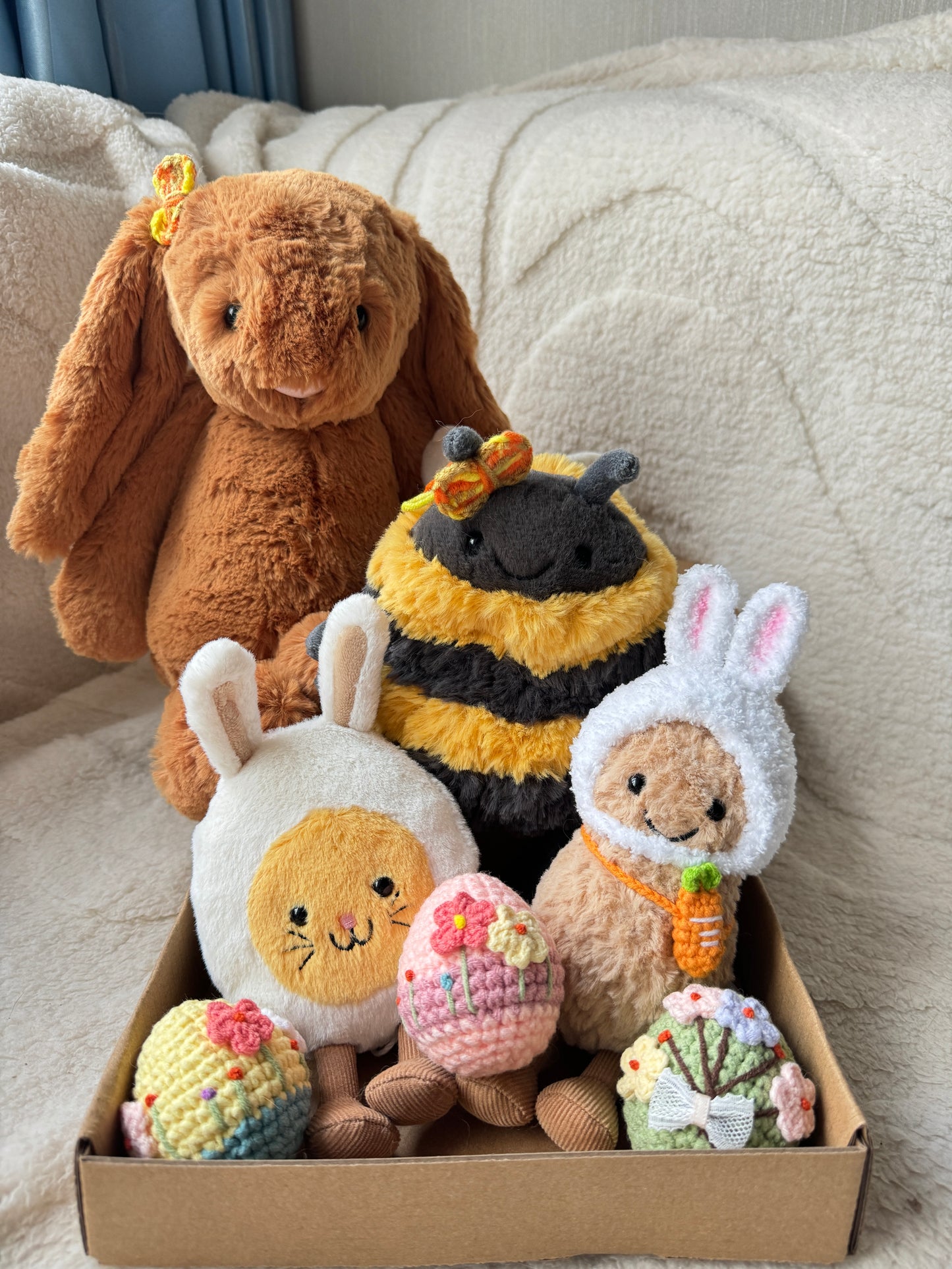 A deluxe handmade Easter box set featuring plush toys dressed in spring-themed outfits, including a large Jellycat bunny with a crochet bow, a bee plush with butterfly clips, bunny-themed plushies, and three embroidered crochet Easter eggs, displayed in a kraft gift box on a soft cream blanket