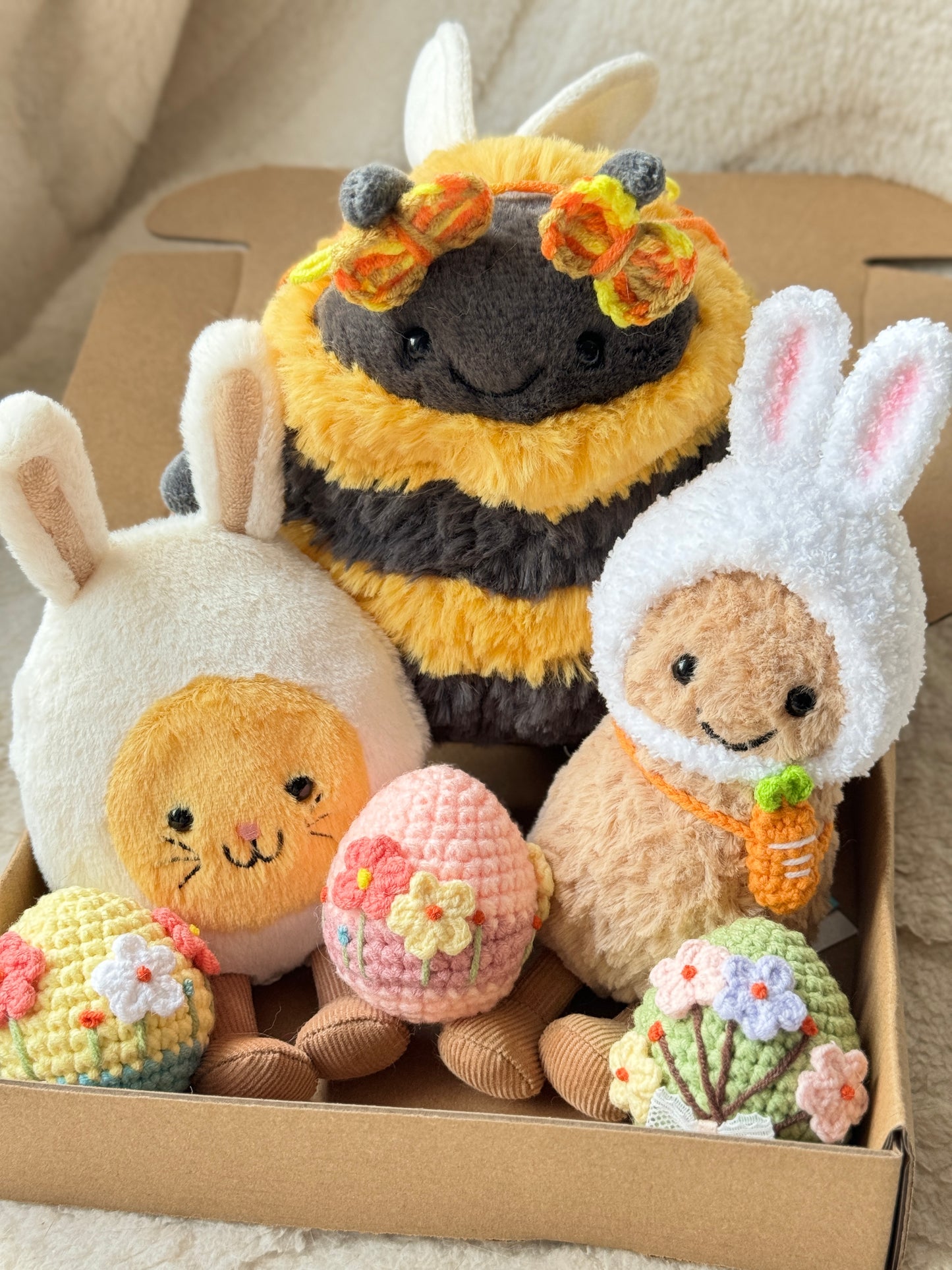 Close-up of three Jellycat plush toys styled for Easter with crochet accessories, including a bunny-hooded plush, a bee plush with butterfly clips, and three pastel crochet Easter eggs decorated with flowers, arranged neatly in a kraft box
