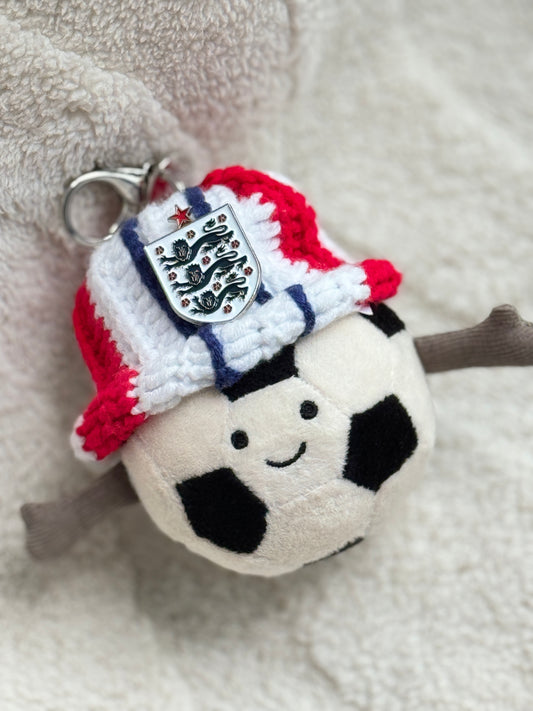 Jellycat Amuseable Sports Football plush wearing a handmade England supporter crochet bucket hat in red, white and blue with an England Three Lions badge attached. Cozy football fan aesthetic inspired by World Cup and England football culture.