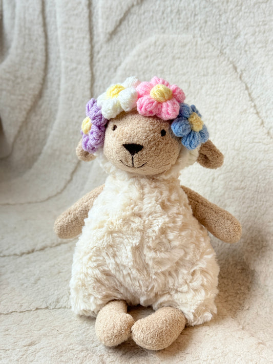 A soft, plush Jellycat sheep wearing a handmade crochet floral headband in pastel colors—white, pink, blue, and lavender flowers with yellow centers. The headband adds a dreamy, fairycore touch to the plush.
