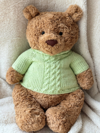 Jellycat Bartholomew Bear wearing a handmade green cable knit sweater, styled on a soft cream blanket with a cozy plush look.