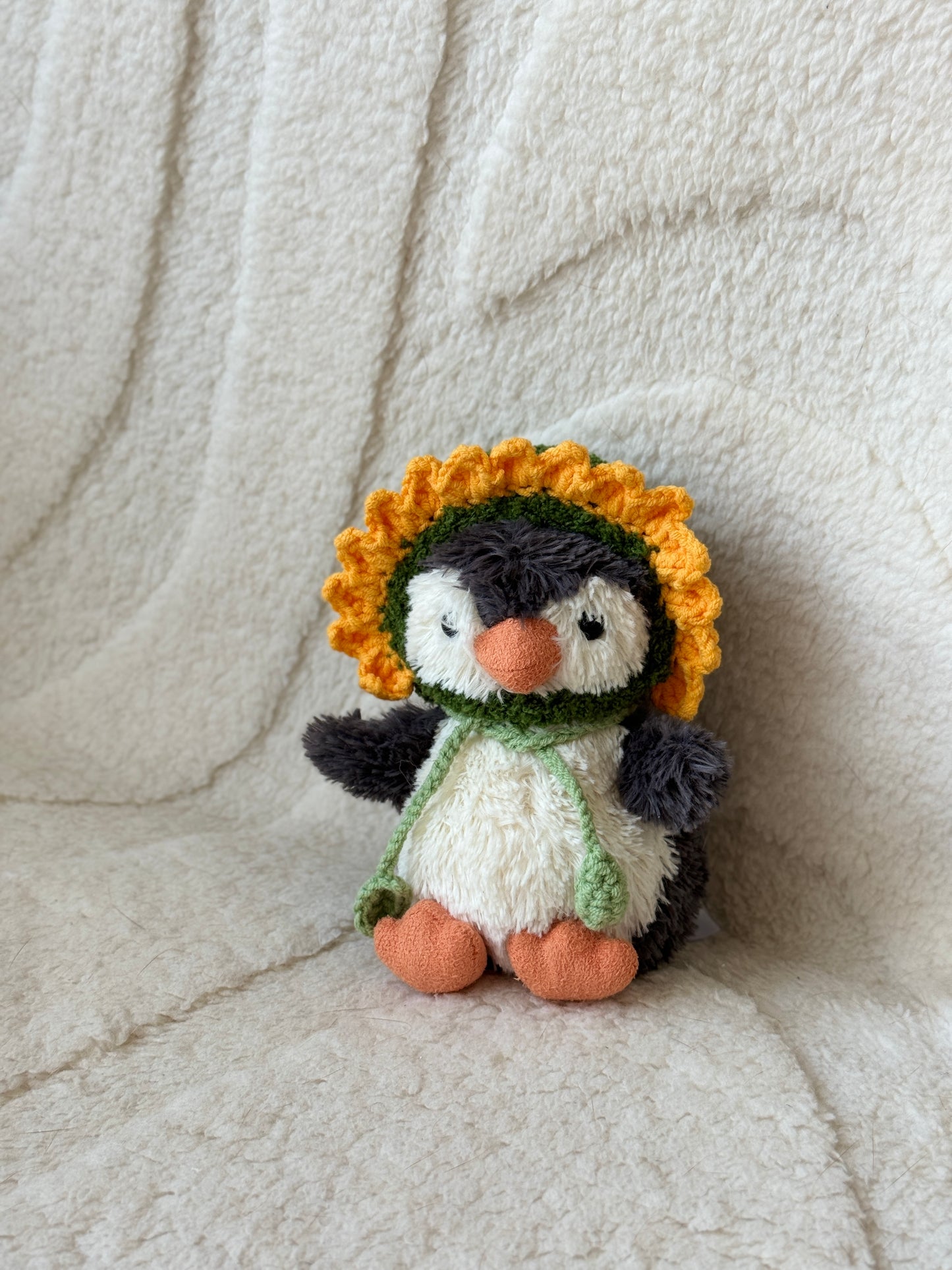 Jellycat Little Penguin plush wearing a handmade crochet sunflower bonnet with a yellow petal trim and green chin ties, sitting against a soft cream background.

