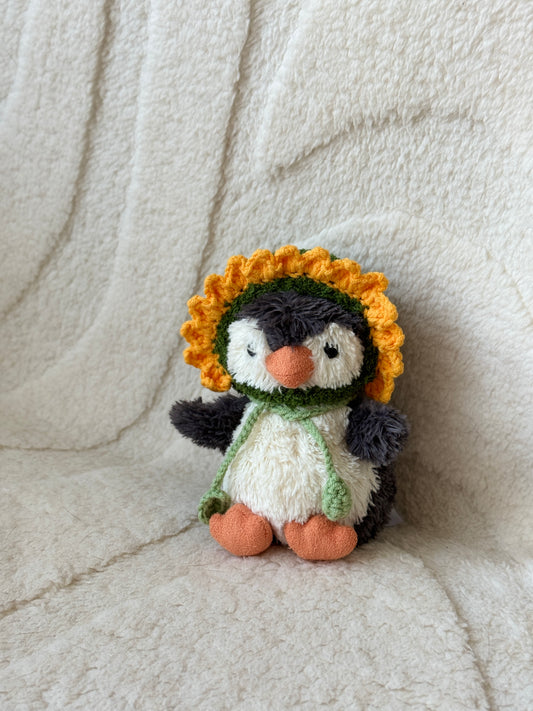 Jellycat Little Penguin plush wearing a handmade crochet sunflower bonnet with a yellow petal trim and green chin ties, sitting against a soft cream background.

