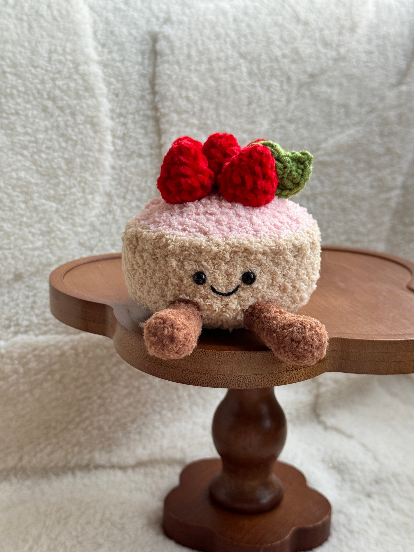 crochet plush of Jellycat Fleurette Tarte aux Fraises with red strawberries and smiling face on a wooden dessert stand.