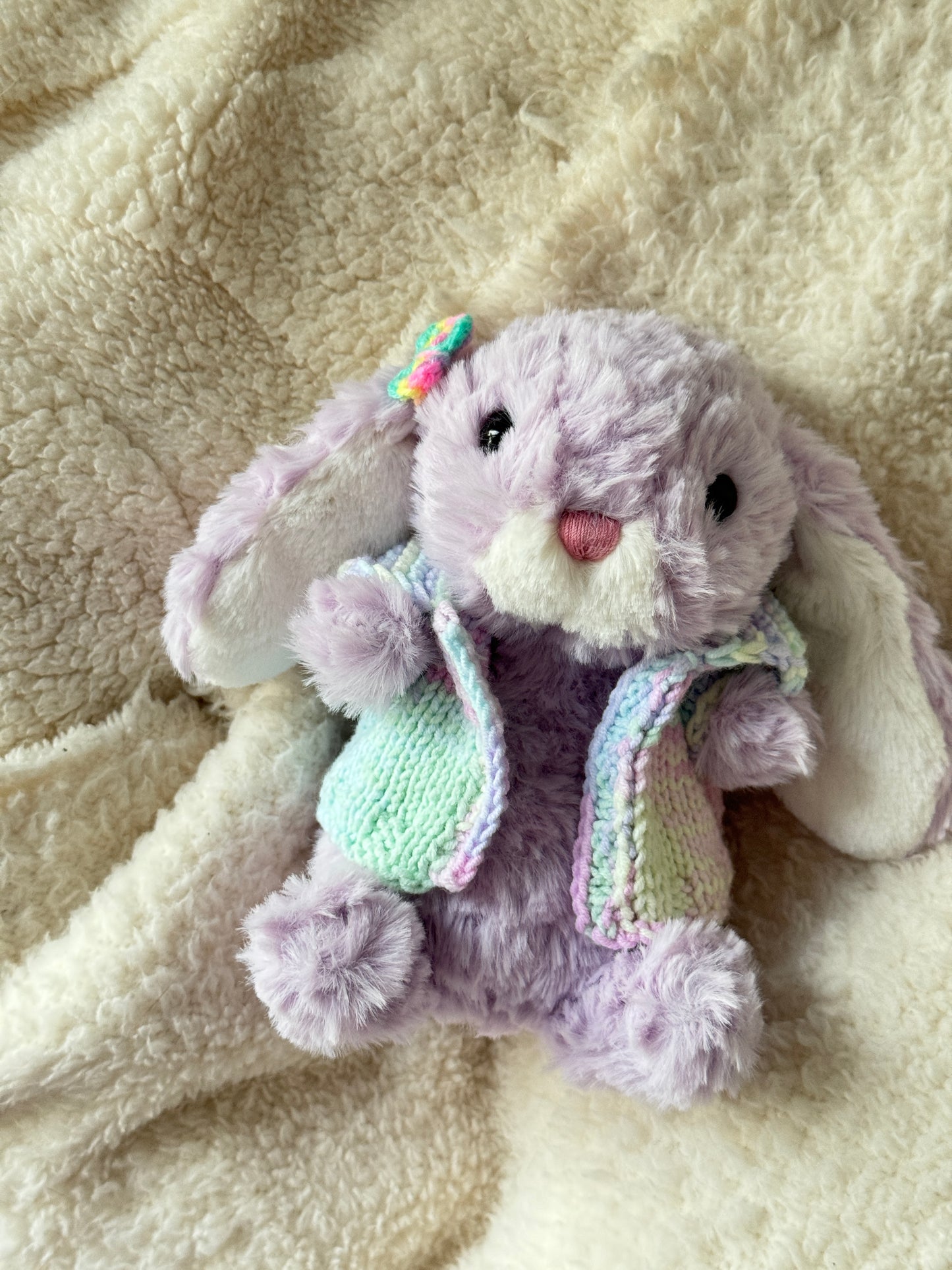 Jellycat Little Bunny 18 cm wearing a handmade pastel knit vest on a cream fleece blanket.