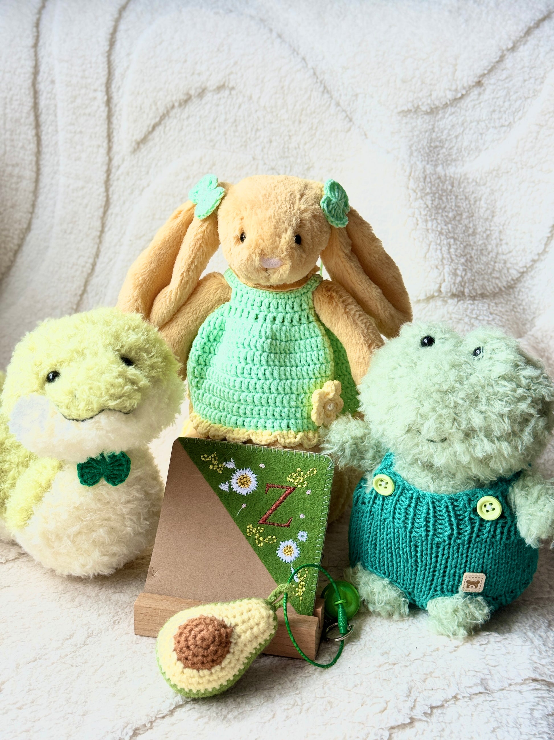 A Jellycat bunny plush dressed in a handmade green crochet dress with matching hair bows. The plush is posed on a soft, cream-colored blanket with other plush friends and festive green accessories.