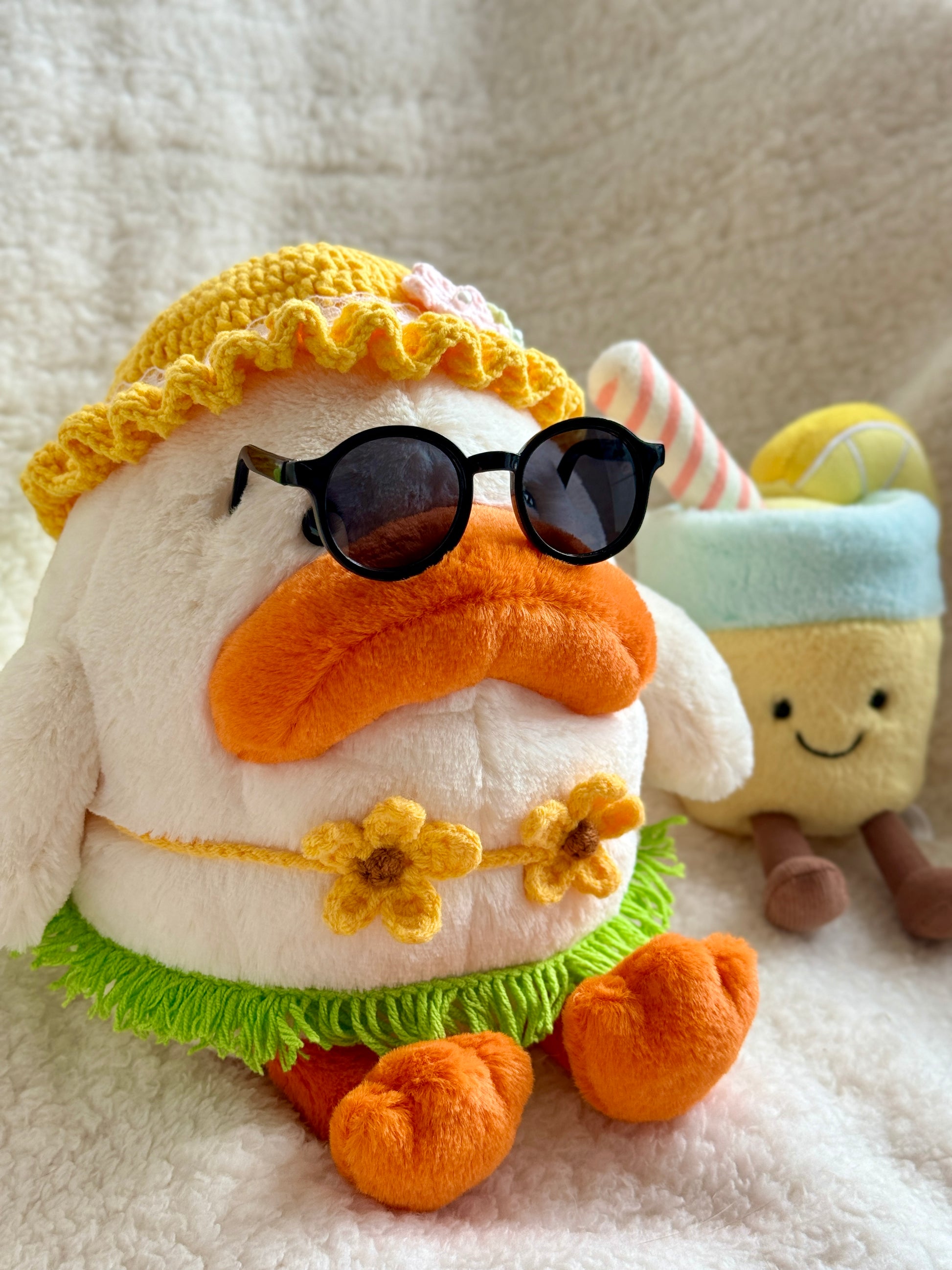 Jellycat Dulia Duck sits upright wearing a handmade yellow sun hat with crochet flowers, black sunglasses, a sunflower bikini top, and a green yarn grass skirt. Two plush drinks — a coconut and a lemonade — are placed on either side of her.

